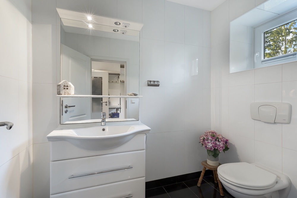 Modernes Badezimmer mit Waschbecken, Spiegel, Toilette und einem Fenster. Eine Vase mit Blumen steht neben der Toilette.