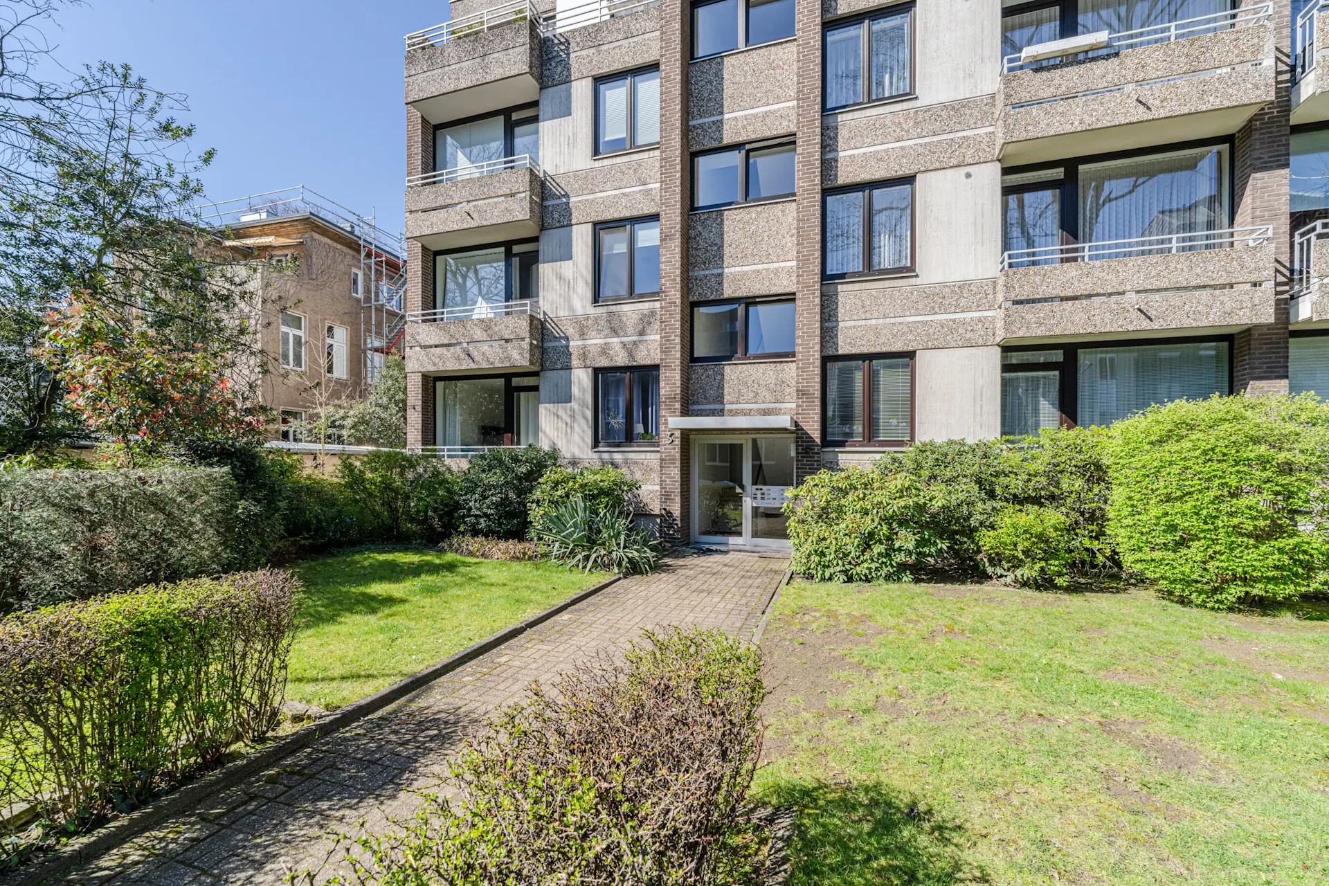 Mehrfamilienhaus mit Balkonen, Garten und gepflastertem Weg zum Eingang bei Sonnenschein.