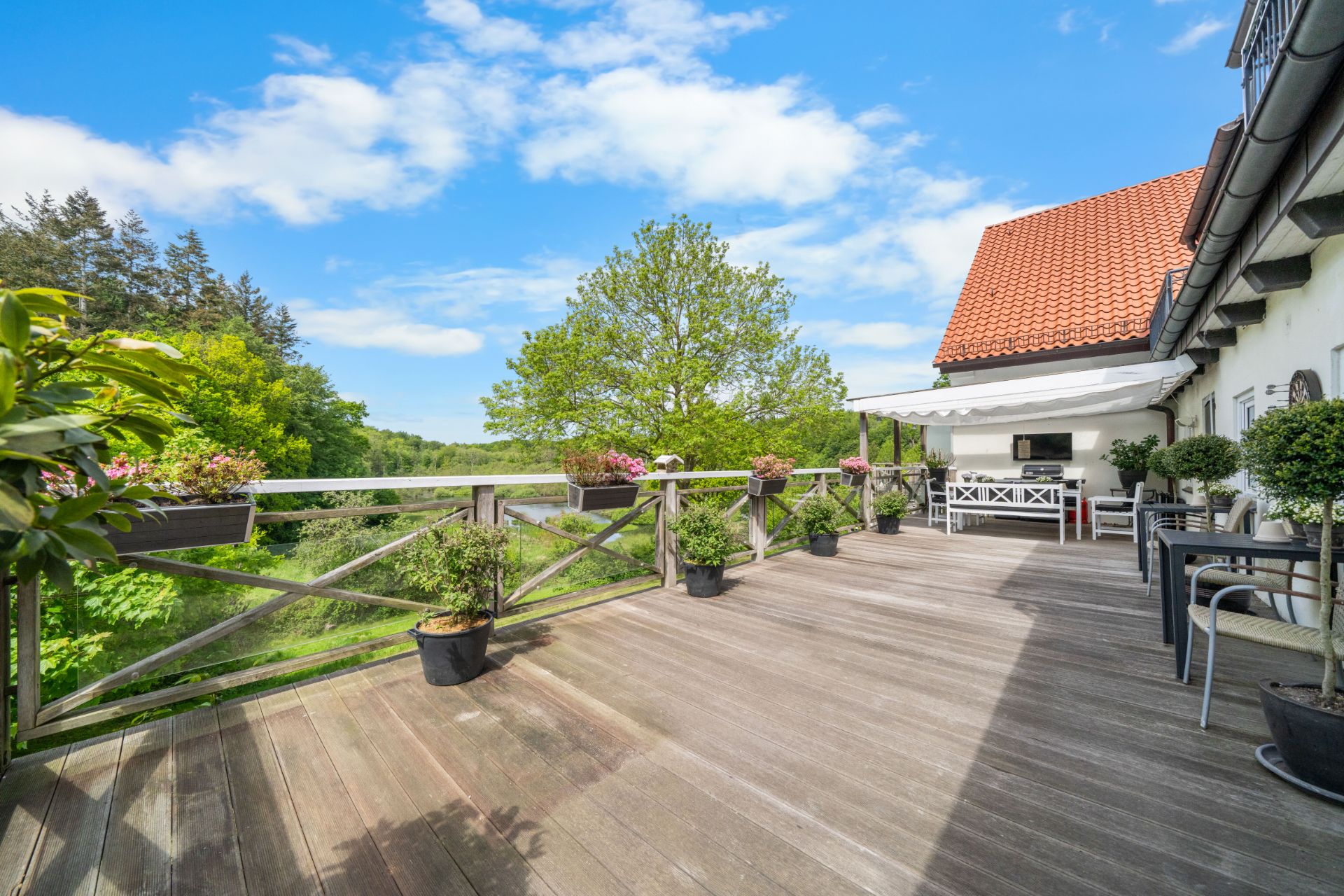 Große Holzterrasse mit Blumentöpfen, Gartenmöbeln und Blick auf grüne Bäume.