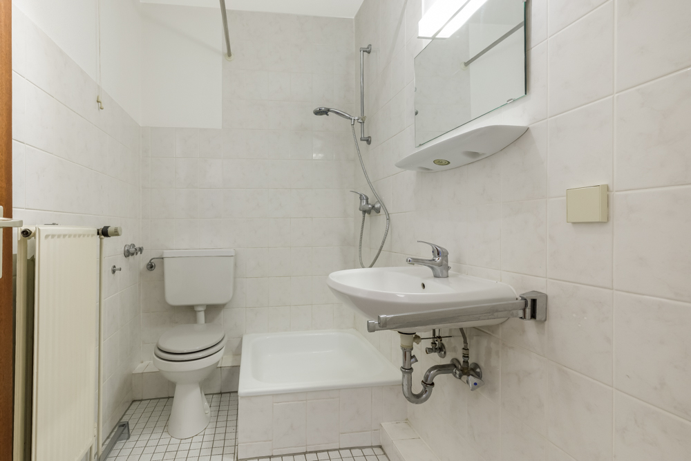 Kleines Badezimmer mit Toilette, Dusche, Waschbecken und Spiegel, gefliest in Weiß.