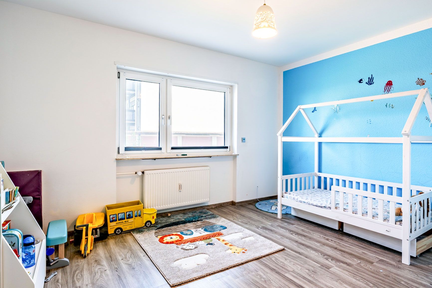 Helles Kinderzimmer mit Teppich, Spielzeug, Bücherregal und weißem Kinderbett vor blauer Wand.