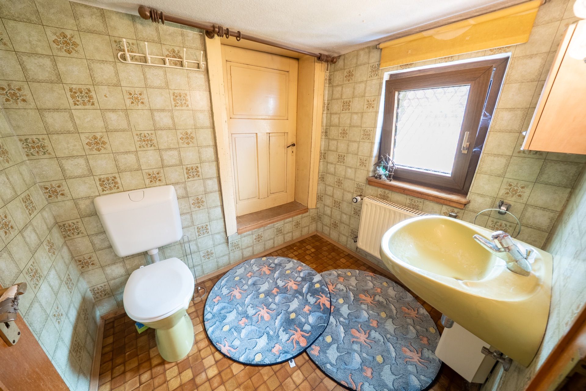 Kleines Badezimmer mit WC, gelbem Waschbecken, Fenster und zwei runden Teppichen auf dem Boden.