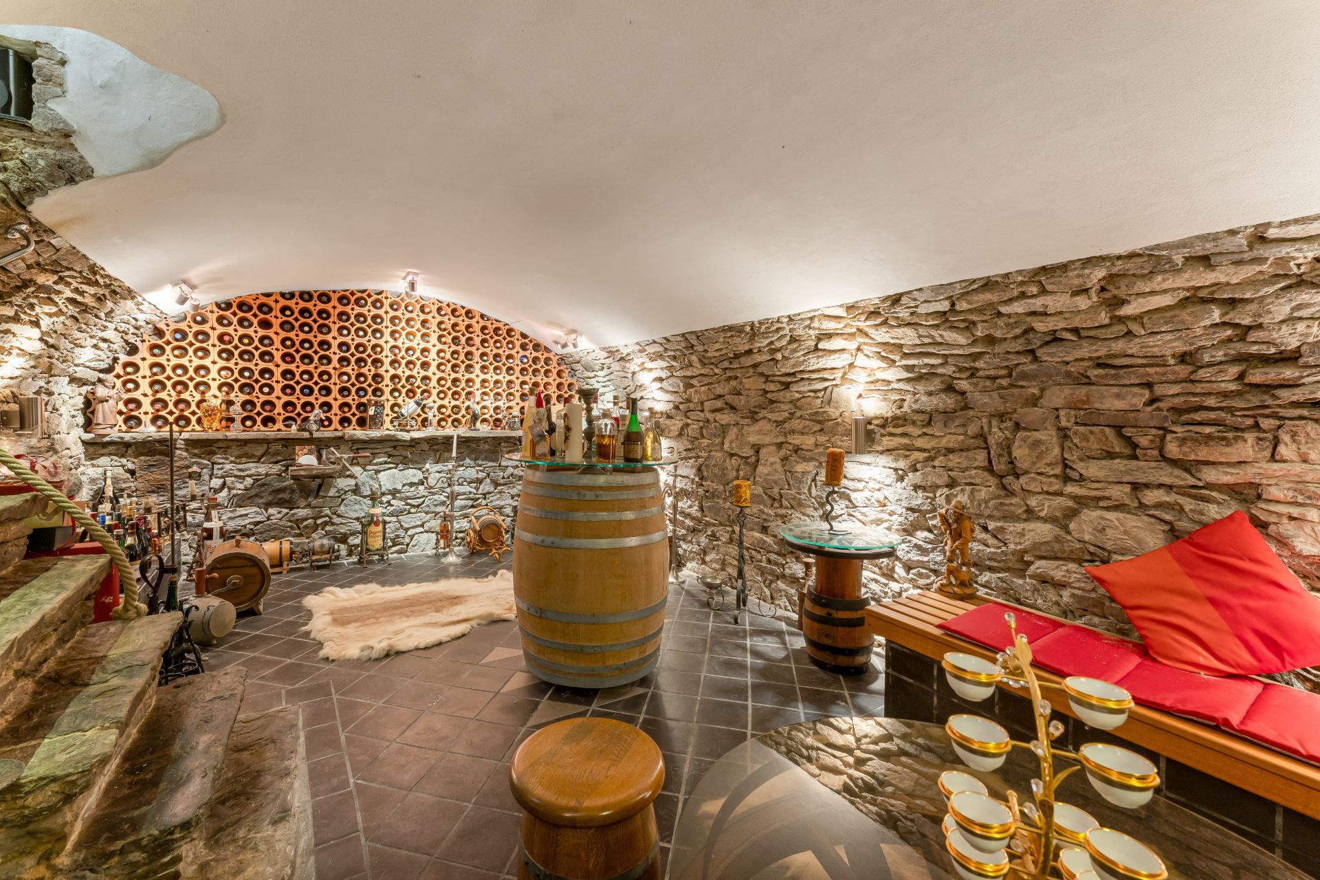 Gemütlicher Weinkeller mit Steinwänden, Weinregalen, Holzmöbeln und einer Sitzbank mit roten Kissen.