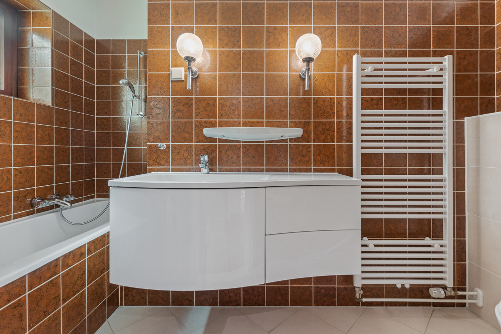 Badezimmer mit braunen Fliesen, weißem Waschbecken, Badewanne und Handtuchheizkörper.