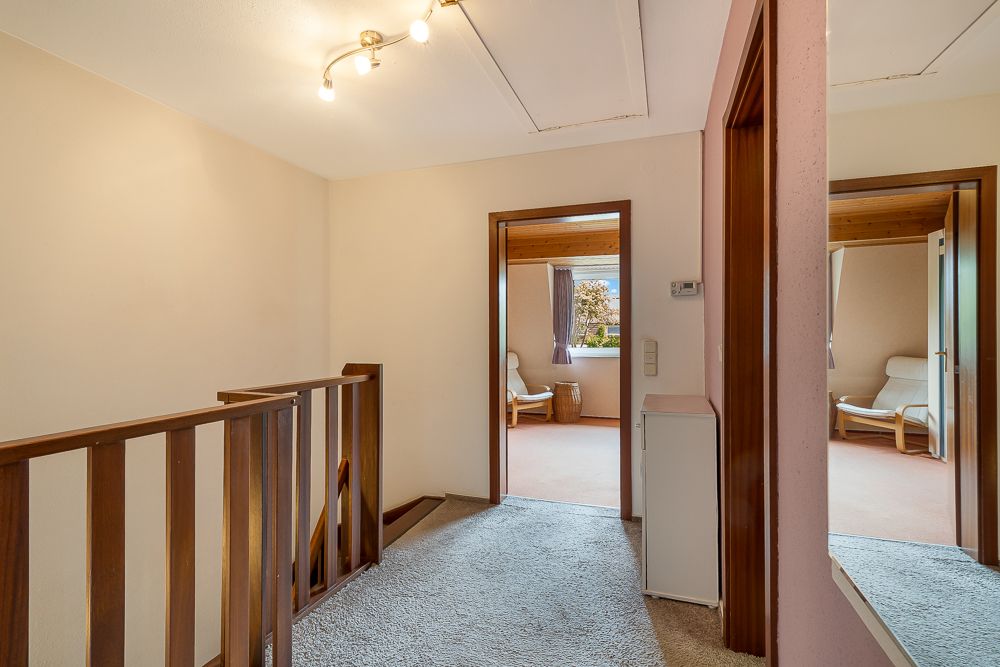 Heller Flur mit Teppichboden, Holztreppe und Zugang zu zwei Zimmern mit Sesseln und Fenstern.