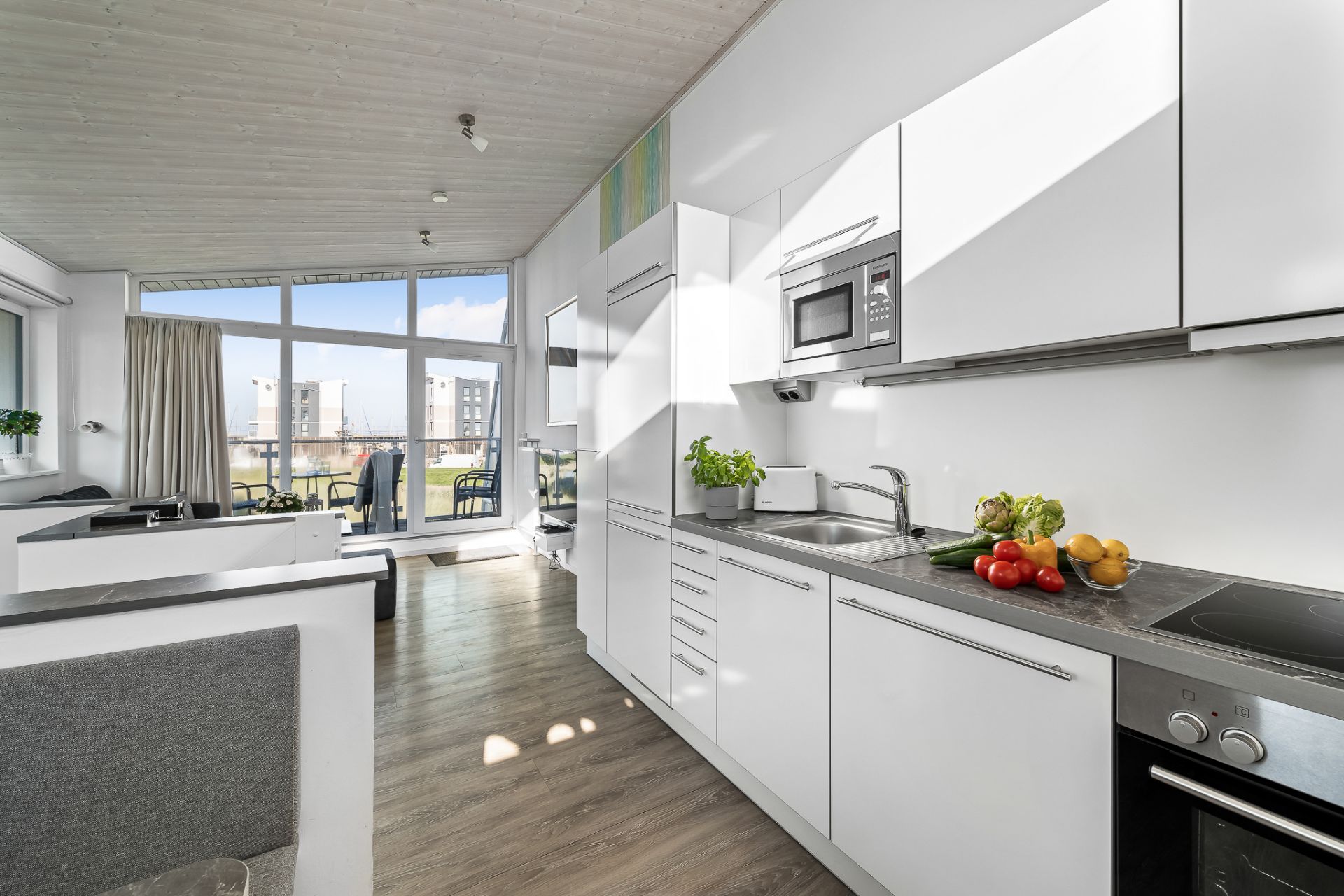 Moderne weiße Küche mit Blick auf ein helles Wohnzimmer und große Fenster.