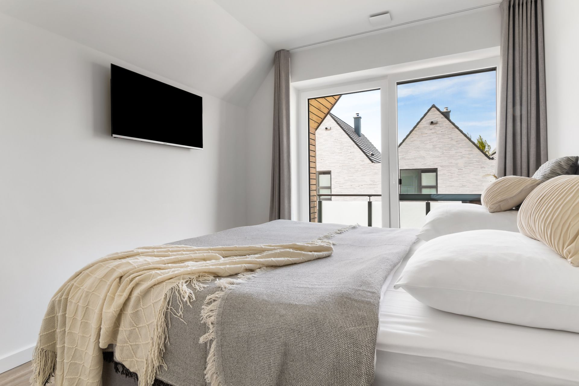 Modernes Schlafzimmer mit Doppelbett, Fernseher an der Wand und großem Fenster mit Blick auf Nachbarhäuser.