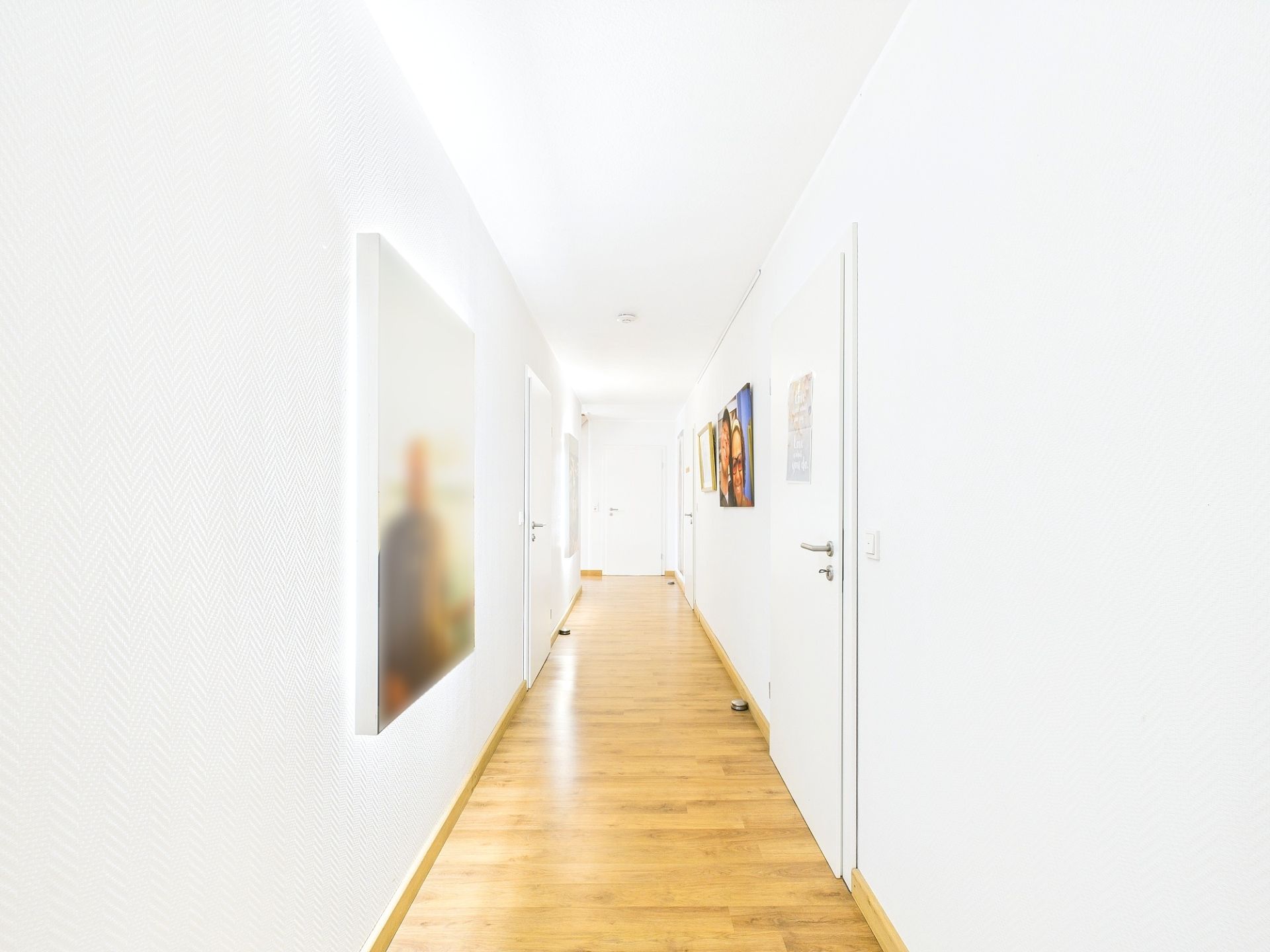 Heller Flur mit Holzboden, weißen Wänden und mehreren Türen, Bilder hängen an den Wänden.