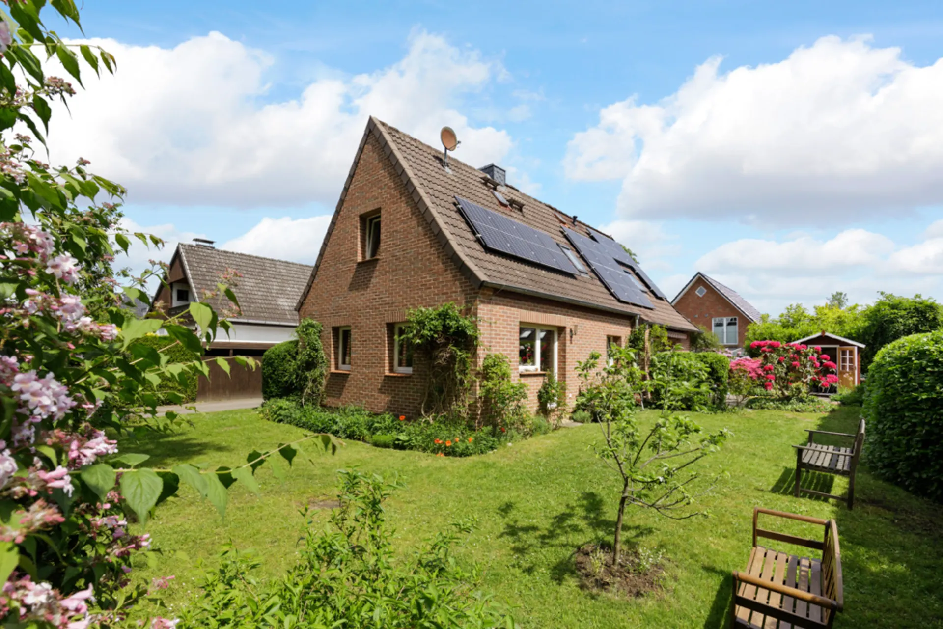 Backsteinwohnhaus mit Solarpaneelen und gepflegtem Garten mit Bänken und Blumen.