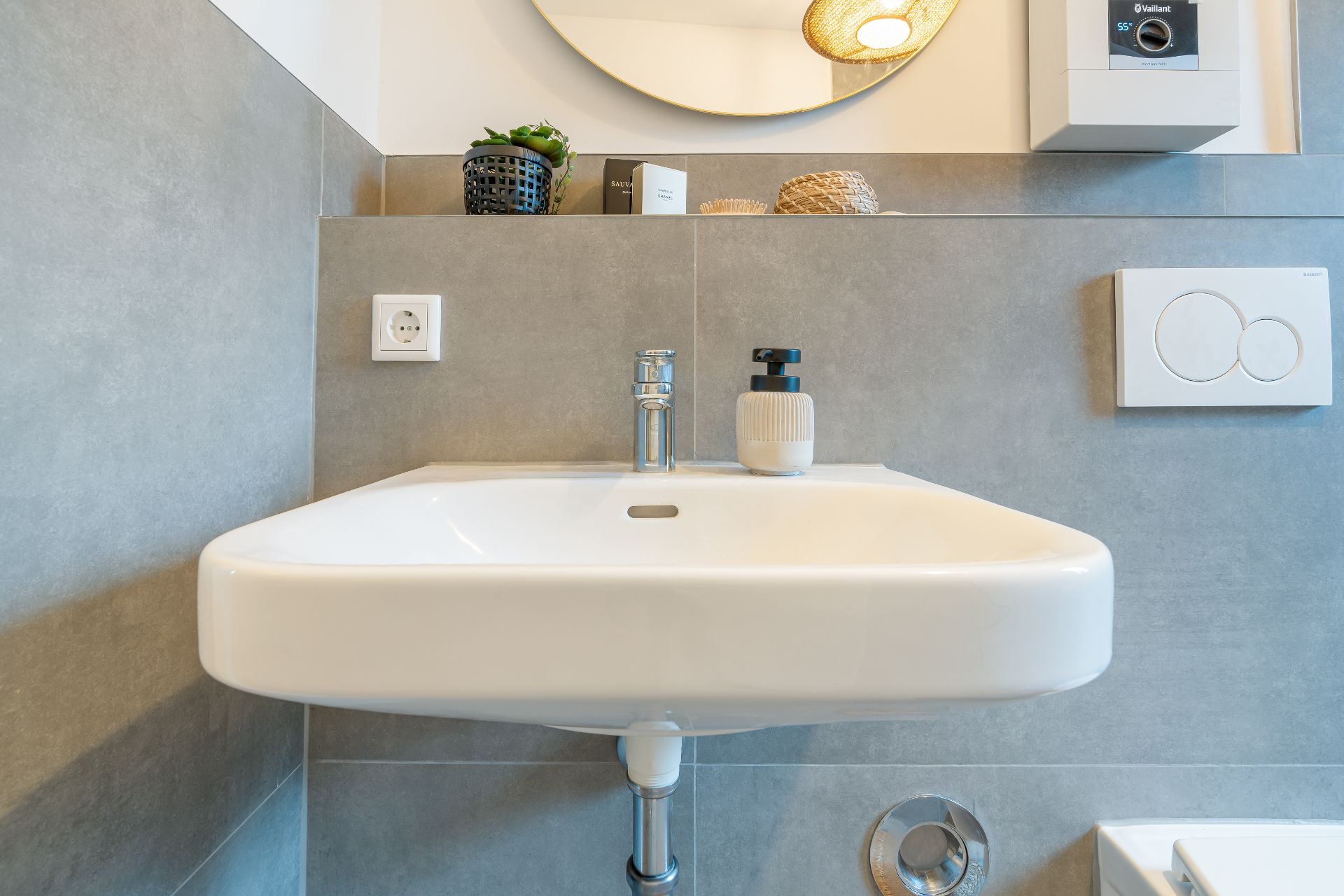 Waschbecken mit Seifenspender, Steckdose und Spülknopf an grauer Wand.