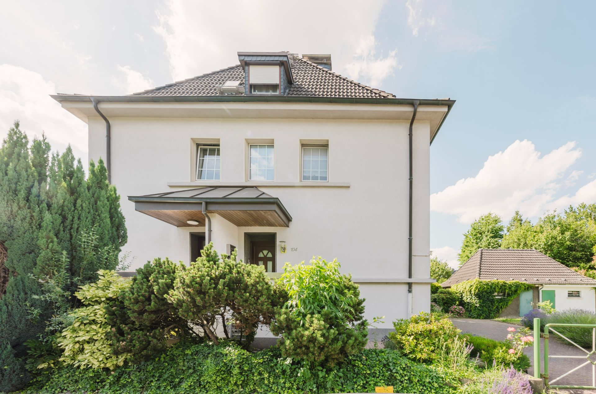 Zweistöckiges Wohnhaus mit Satteldach, Vorgarten und vielen grünen Pflanzen.