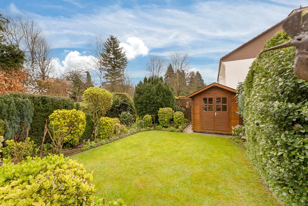 Gepflegter Garten mit Rasen, Sträuchern und einem braunen Gartenhaus im Hintergrund.