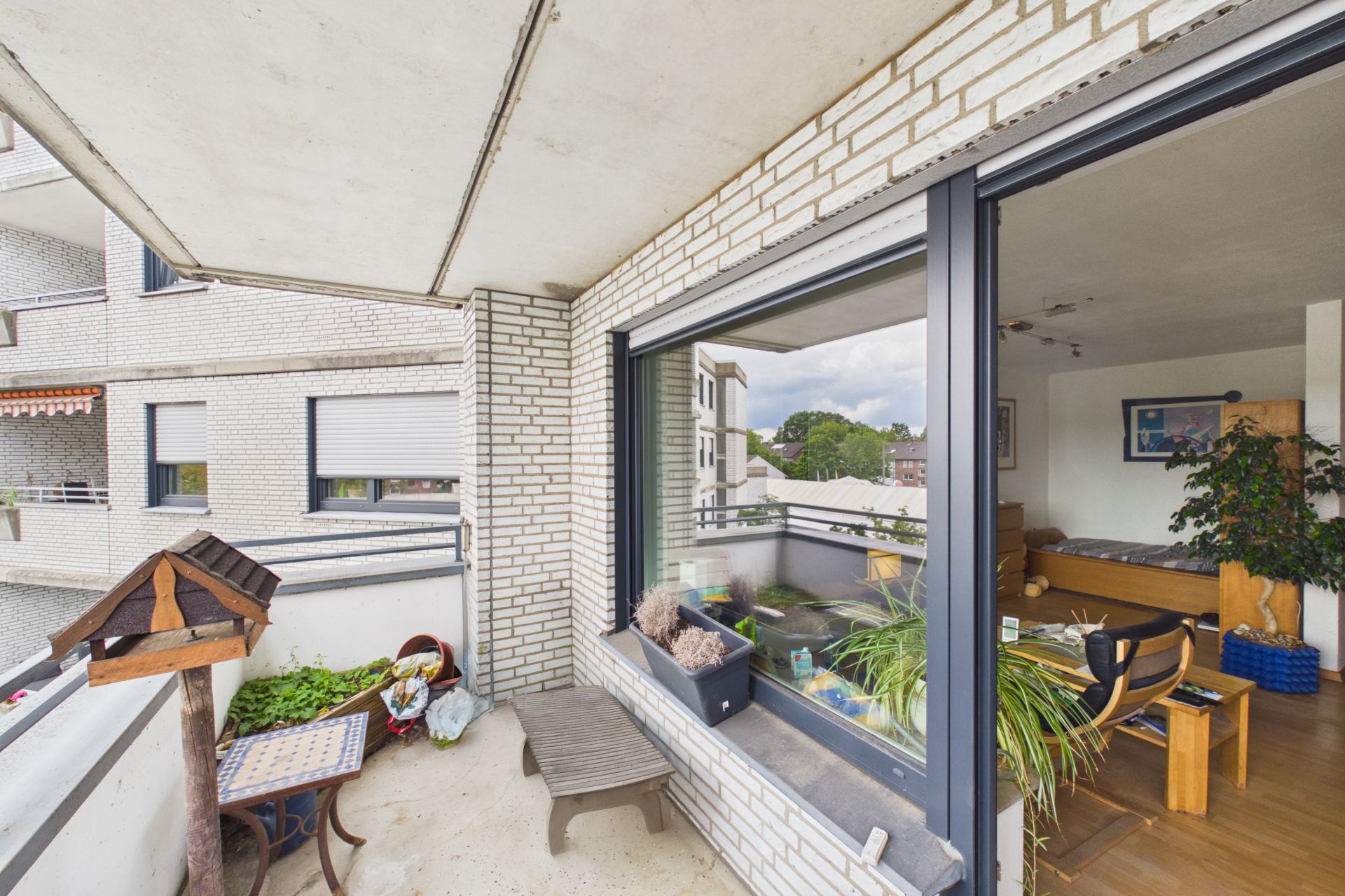 Balkon mit Sitzbank, Vogelhaus, Pflanzen und Blick in ein Wohnzimmer mit Tisch und Sofa.