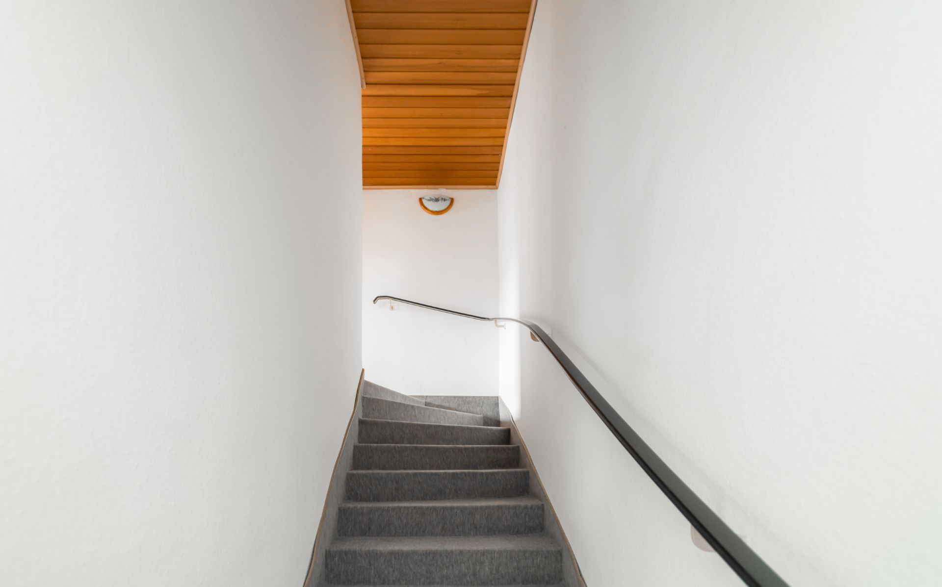 Innenaufnahme einer Treppe mit grauem Belag und Handlauf, weiße Wände, Holzdecke oben.