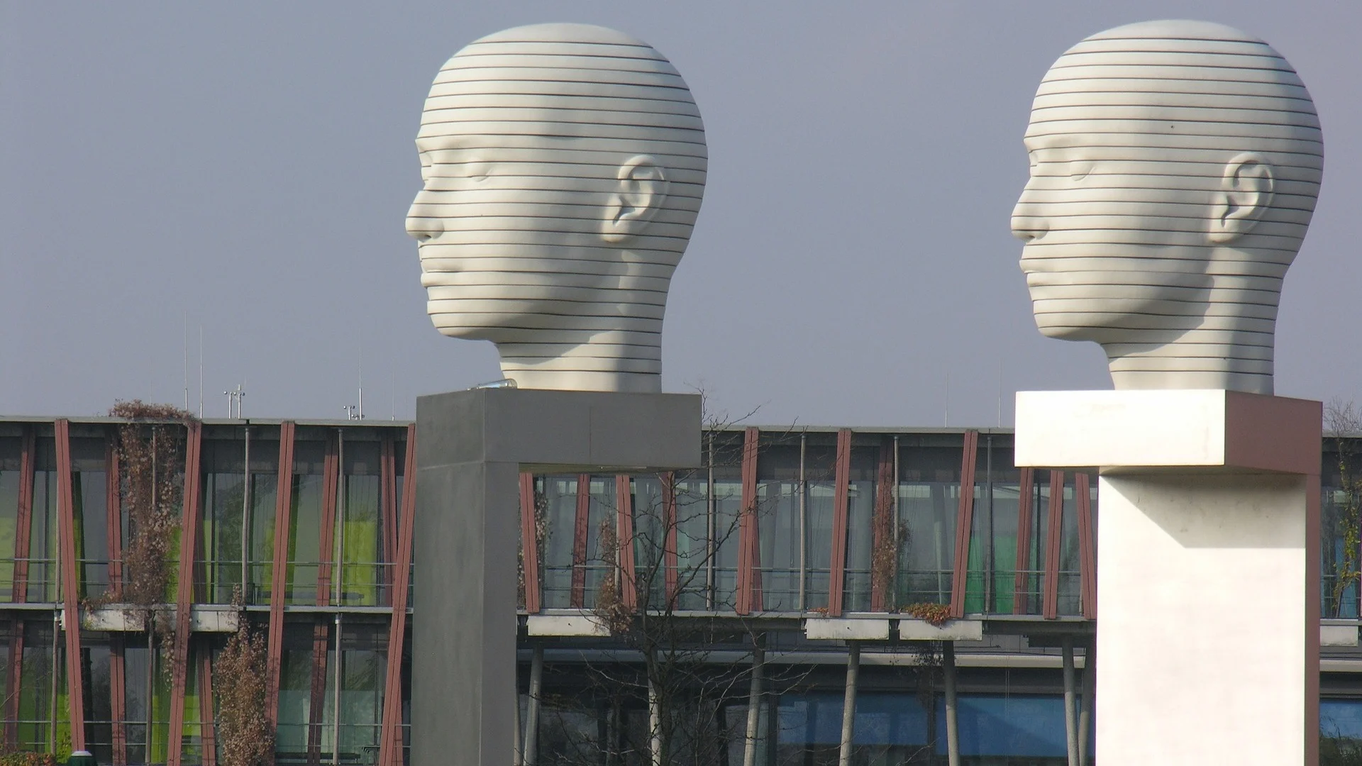 Zwei große, weiße Kopfskulpturen auf rechteckigen Sockeln vor einem modernen Gebäude.