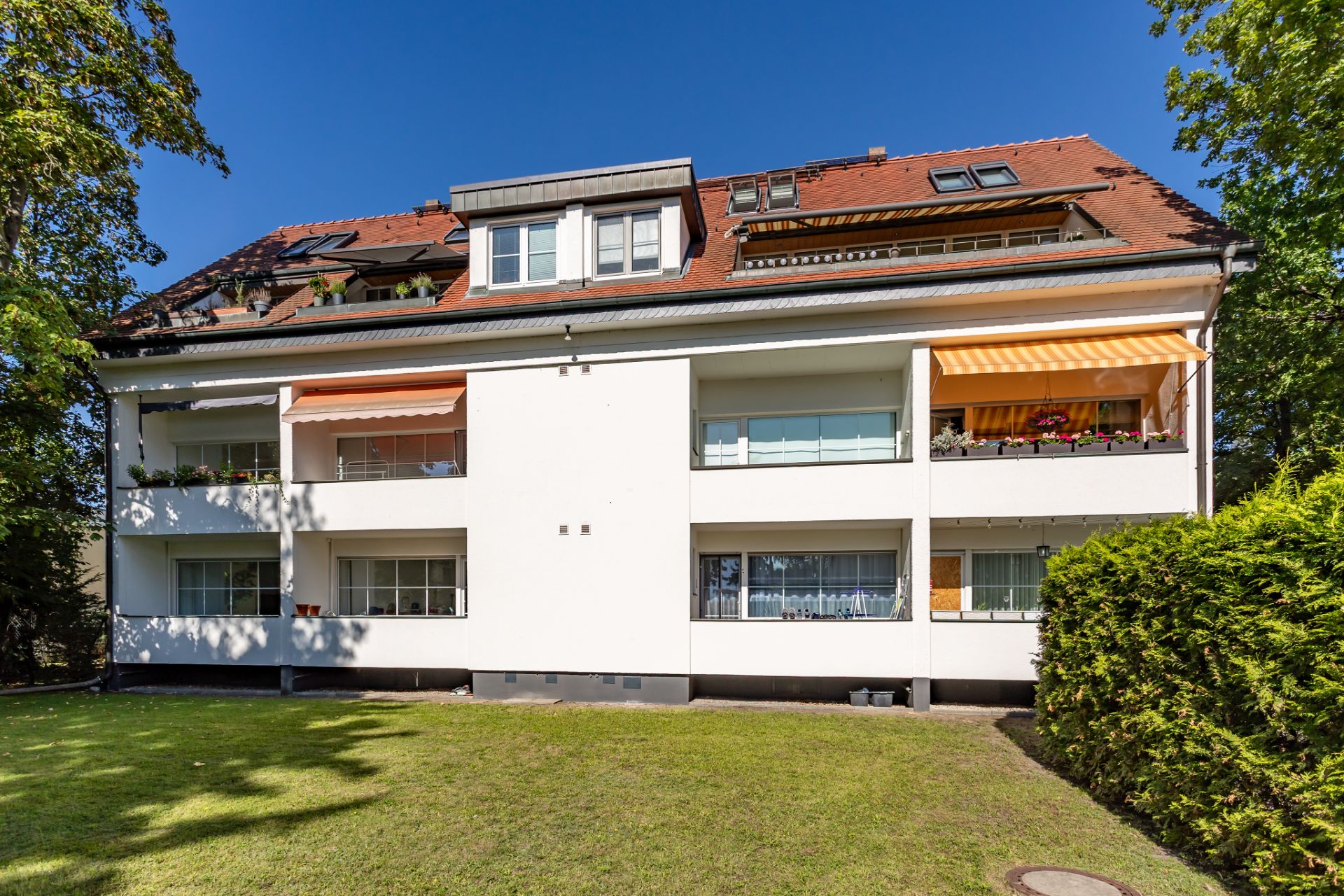 Mehrfamilienhaus mit Balkonen und Markisen, umgeben von grünem Rasen und Bäumen.