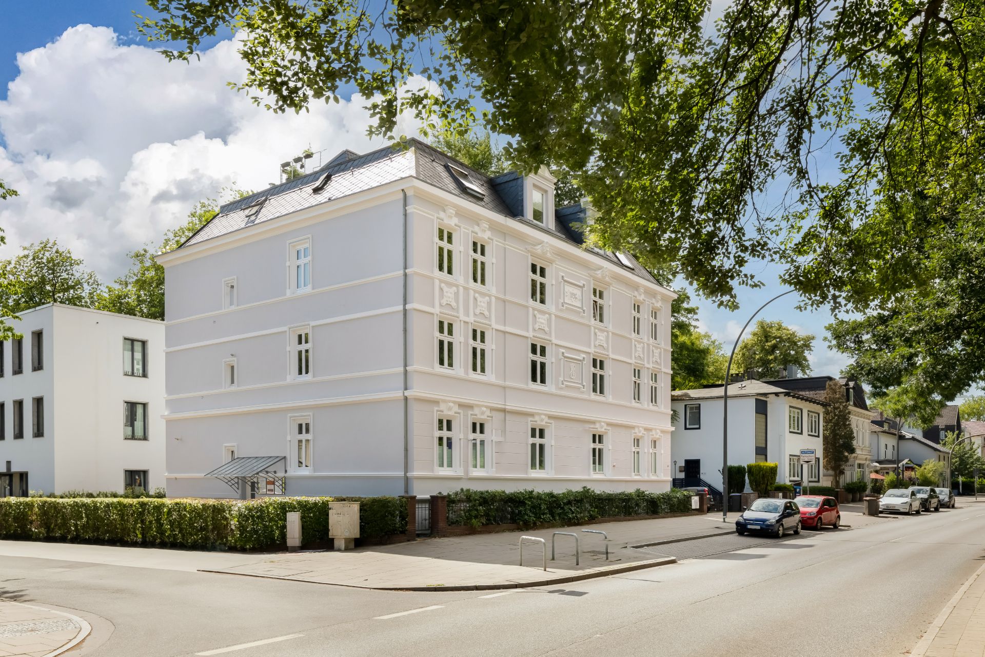Weiße Mehrfamilienhaus an einer ruhigen Wohnstraße mit geparkten Autos und Bäumen.