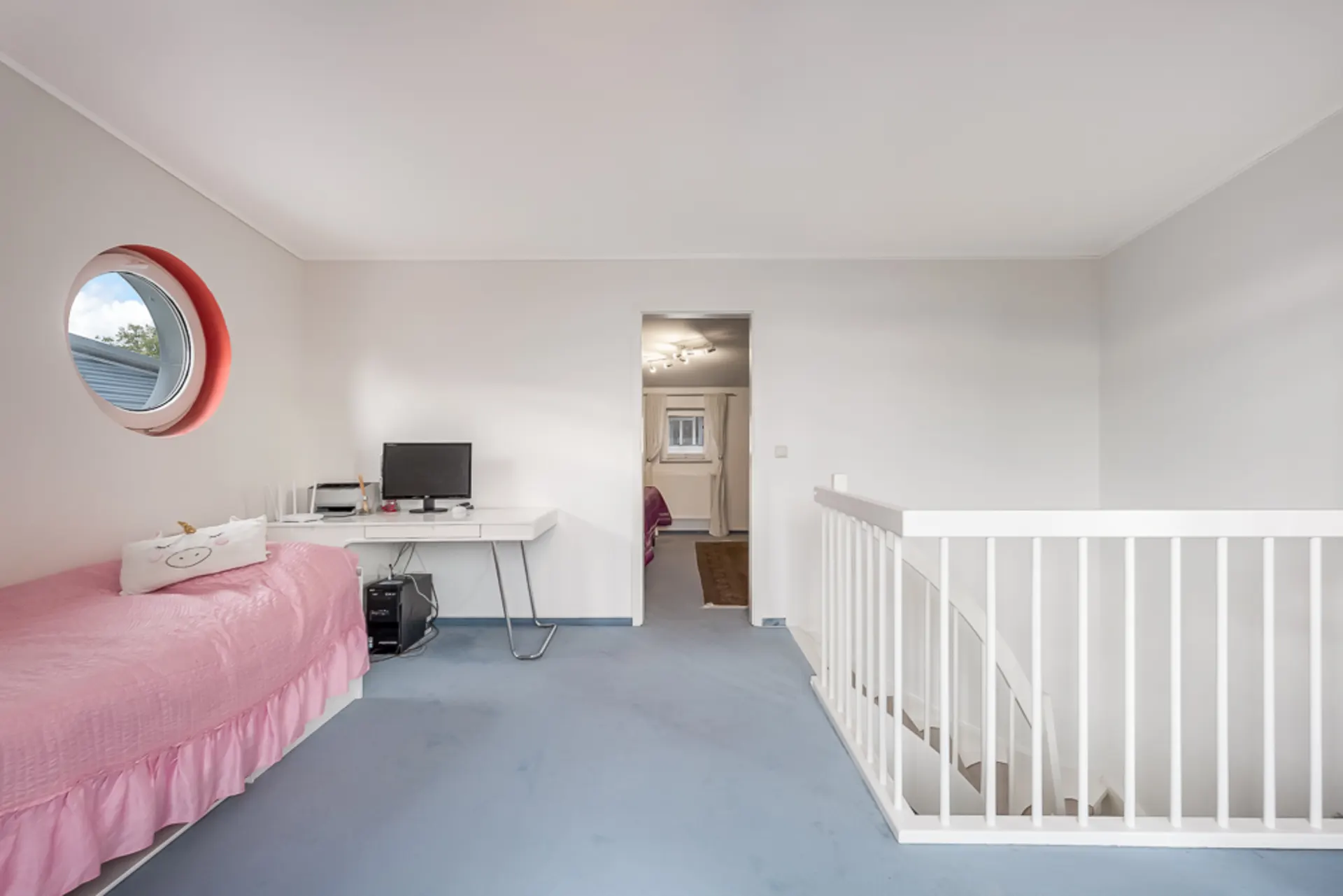 Helles Zimmer mit rundem Fenster, Schreibtisch, Computer, rosafarbenem Bett und Treppengeländer.
