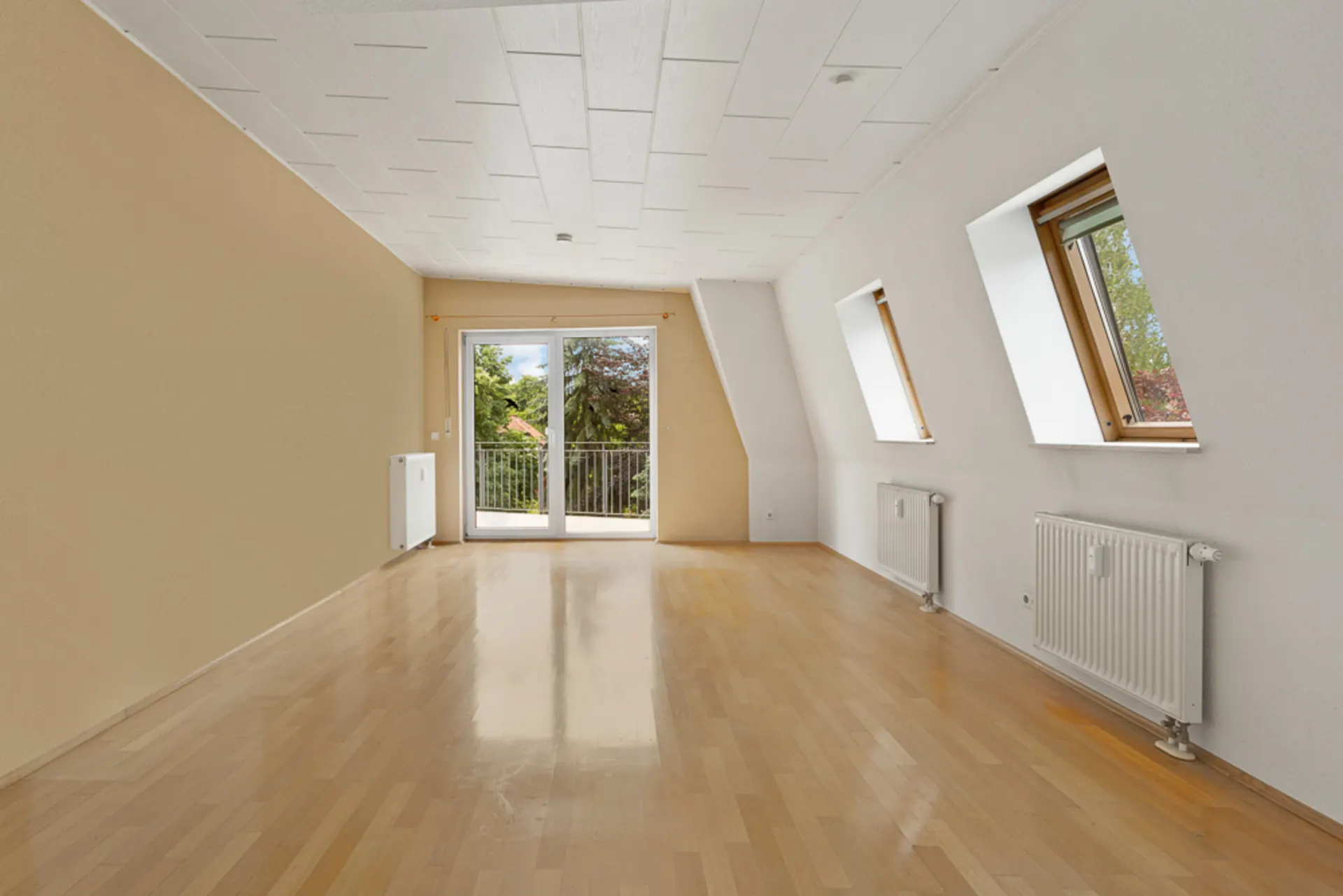 Leeres Zimmer mit Holzboden, drei Fenstern und einer Glastür zum Balkon.