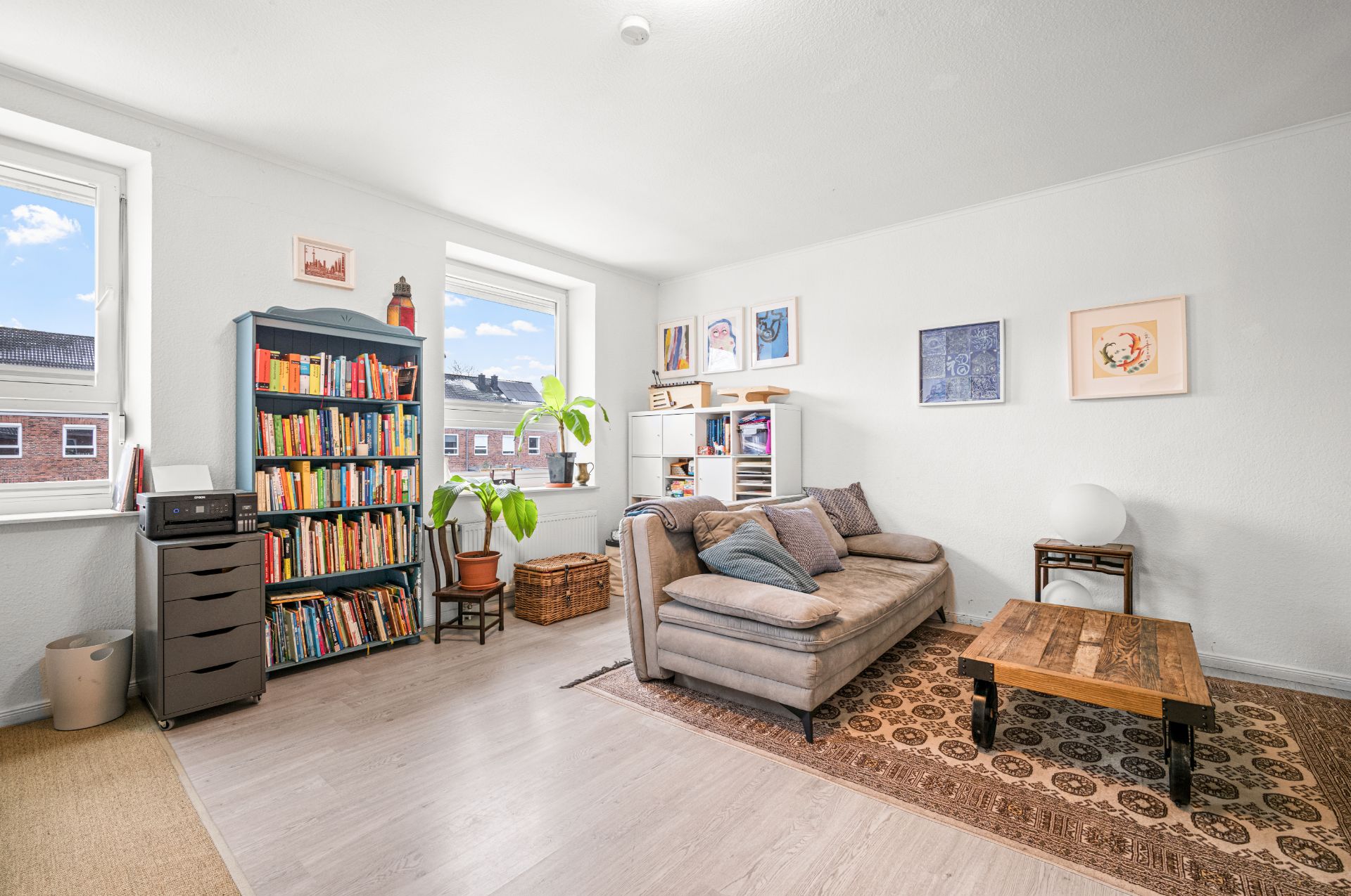 Helles Wohnzimmer mit Sofa, Couchtisch, Bücherregalen, Pflanze und zwei Fenstern.