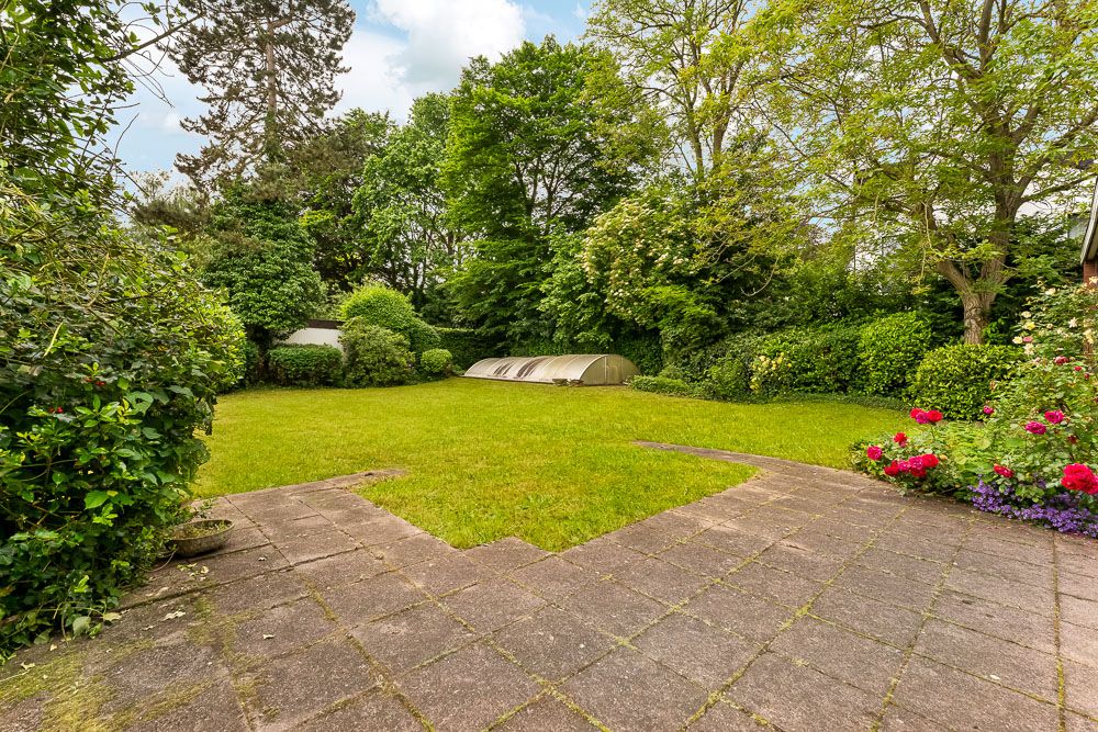 Gepflasterte Terrasse mit Blick auf den grünen Garten, Bäume und Blumenbeete, Rasenfläche in der Mitte.