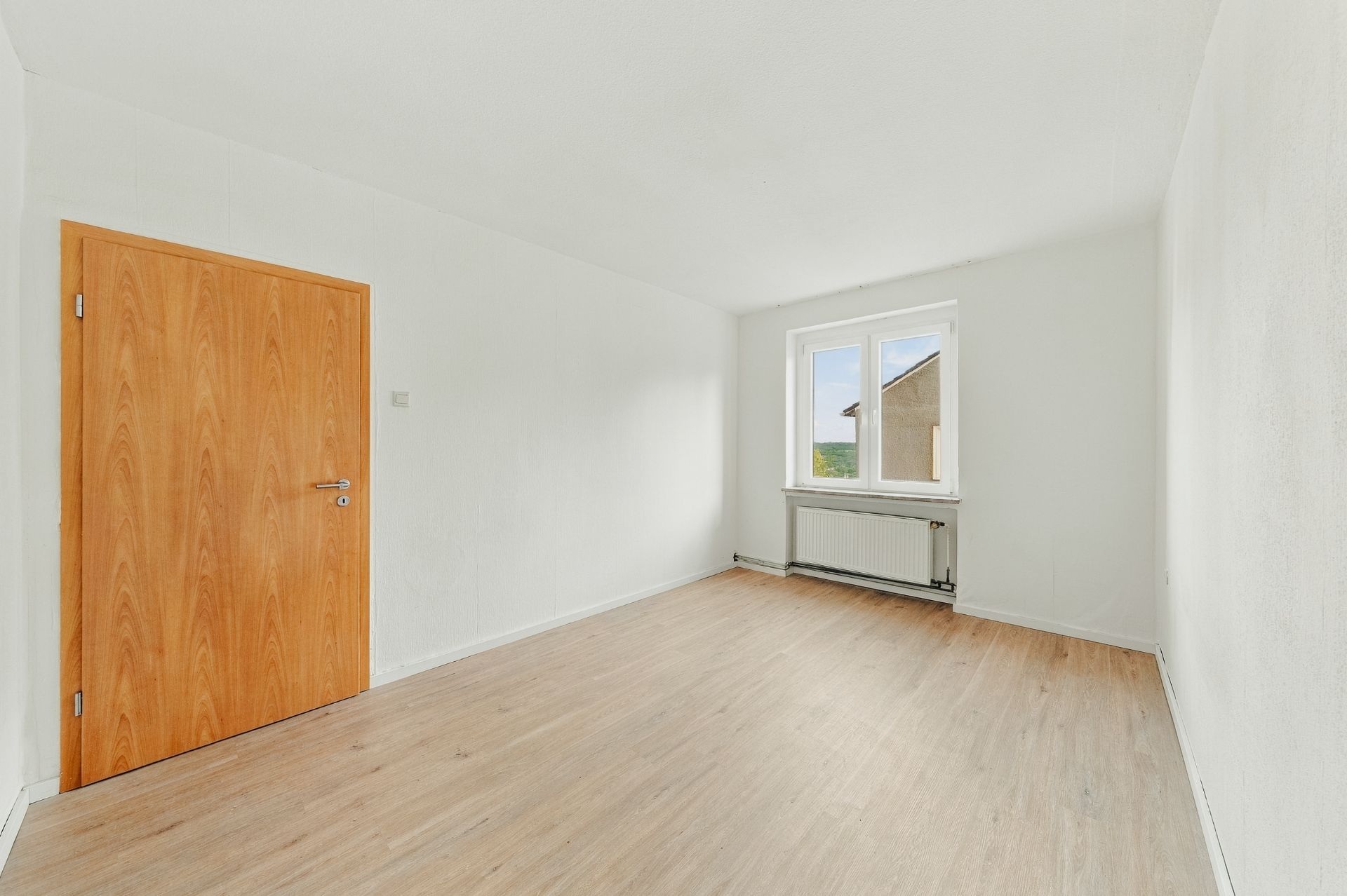 Leeres Zimmer mit weiß gestrichenen Wänden, Holztür, Fenster und Heizkörper.
