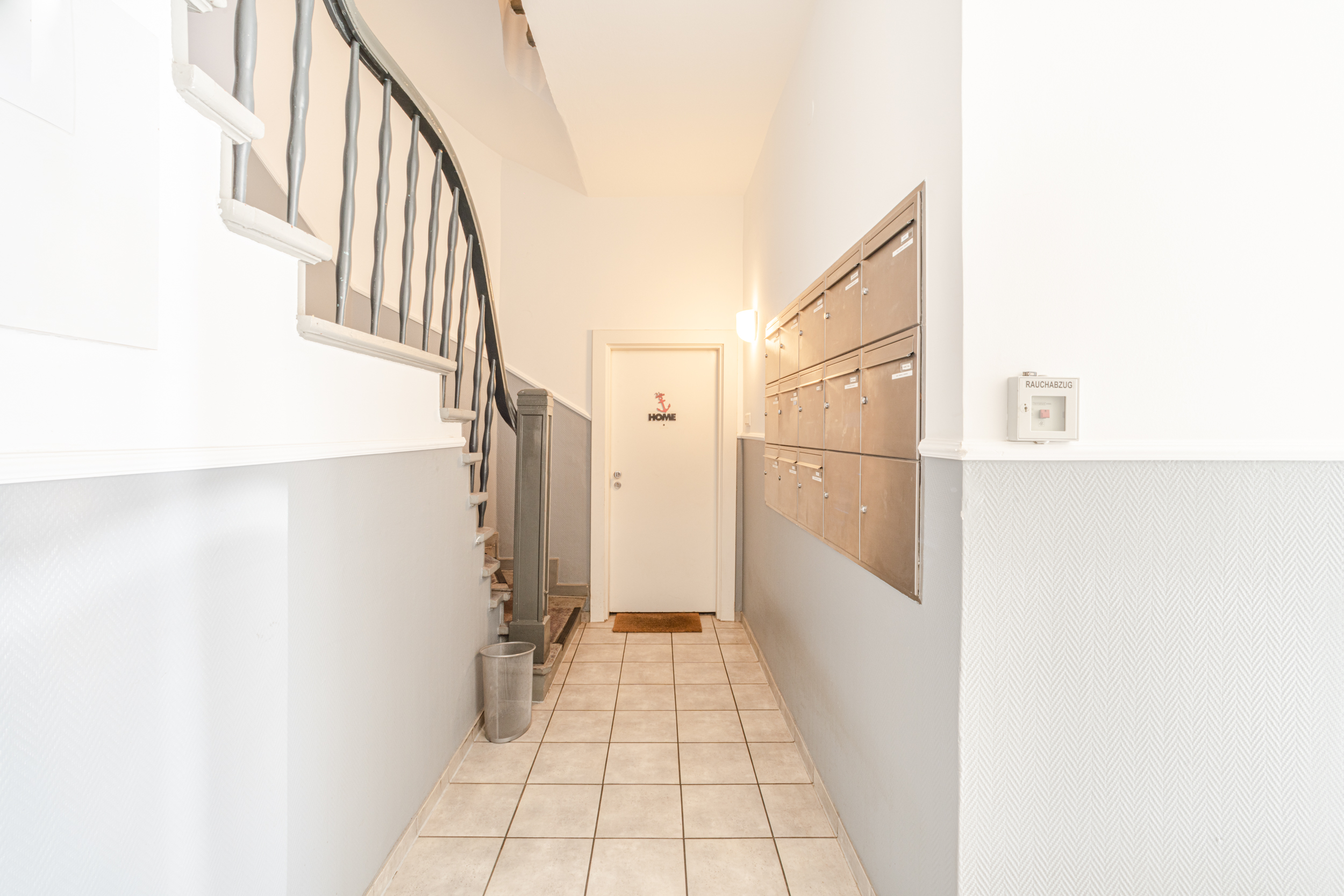 Heller Hausflur mit Treppe, Briefkästen und einer Tür mit dem Schild 'HOME'.