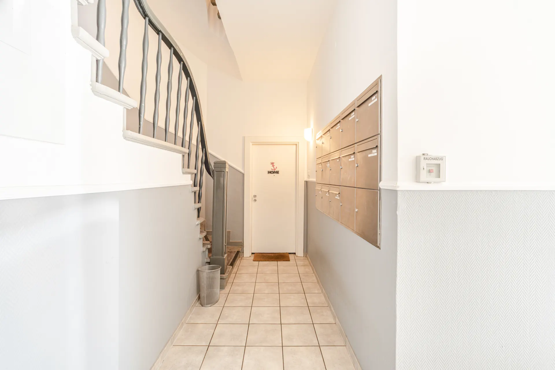 Heller Hausflur mit Treppe, Briefkästen und einer Tür mit dem Schild 'HOME'.