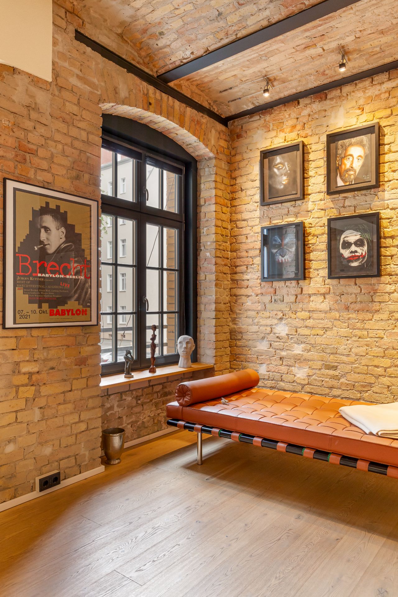 Zimmer mit Backsteinwand, Liege, großen Fenstern und gerahmter Kunst an der Wand.