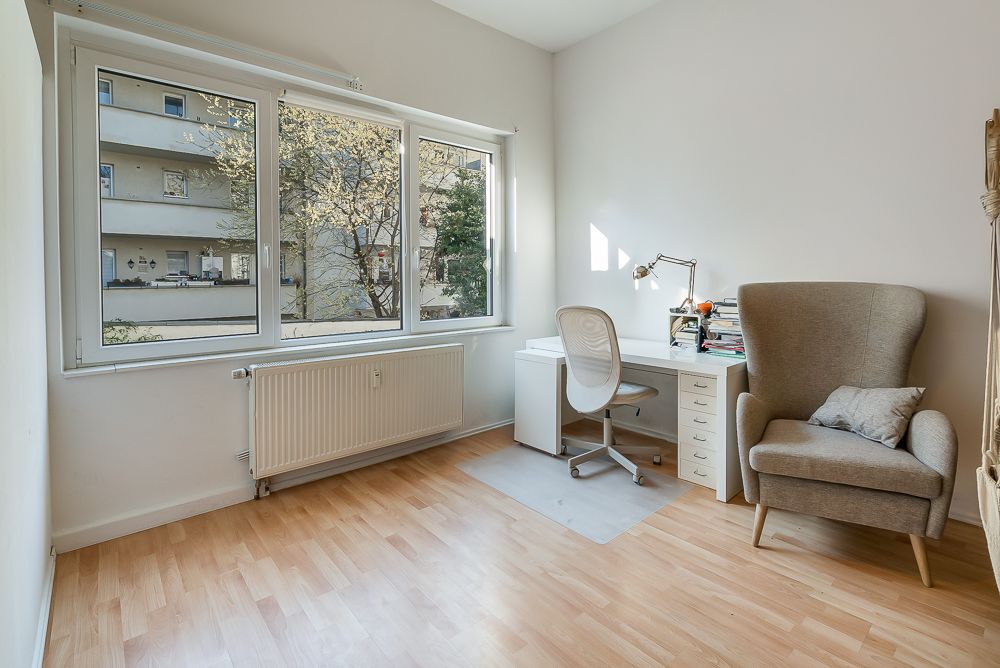 Helles Zimmer mit großem Fenster, Schreibtisch, Stuhl und Sessel auf hellem Holzboden.