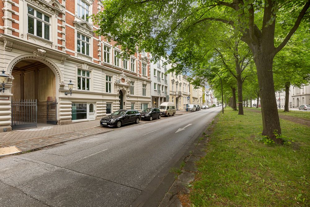 Baumgesäumte Straße mit parkenden Autos vor historischen Wohngebäuden.