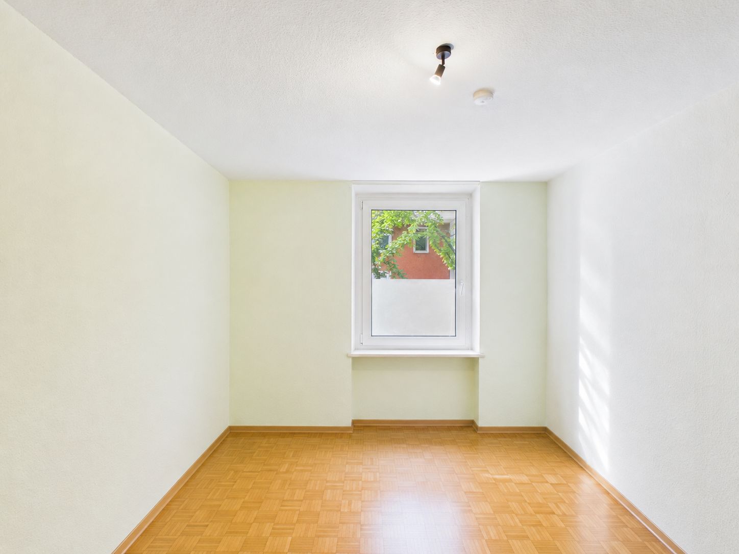 Leerer, heller Raum mit Holzboden und einem Fenster mittig an der Rückwand.