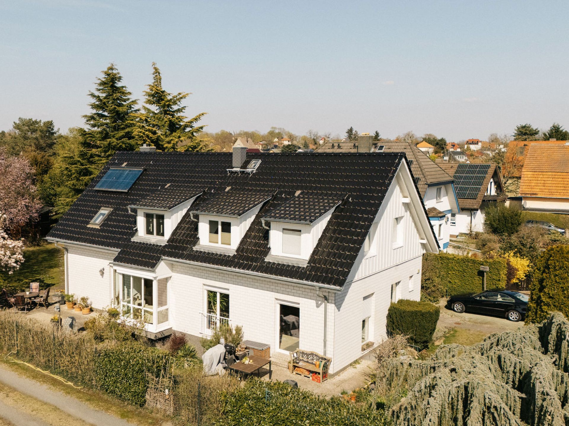 Modernes weißes Wohnhaus mit schwarzem Dach und Terrasse, umgeben von Garten und Bäumen.