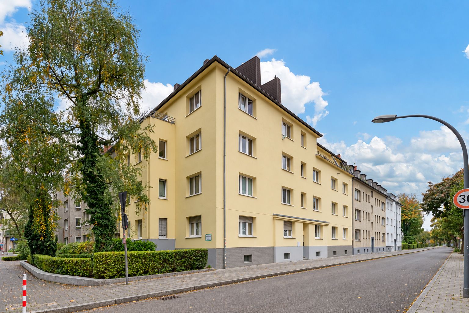 Gelbes Mehrfamilienhaus an einer ruhigen Straße mit Gehweg und Bäumen.