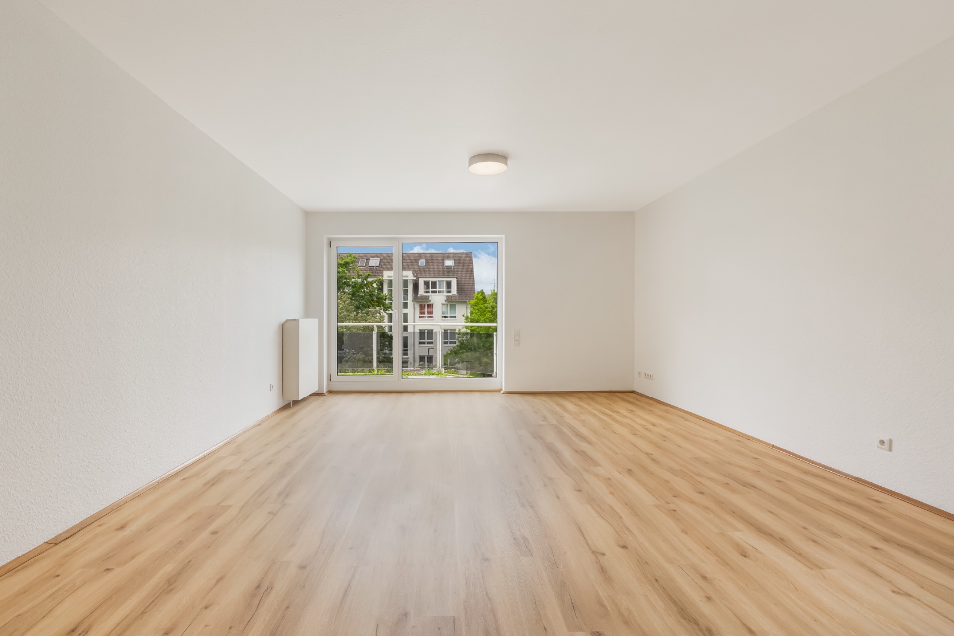 Helles, leeres Zimmer mit Holzboden und großer Fensterfront zu einem Balkon.