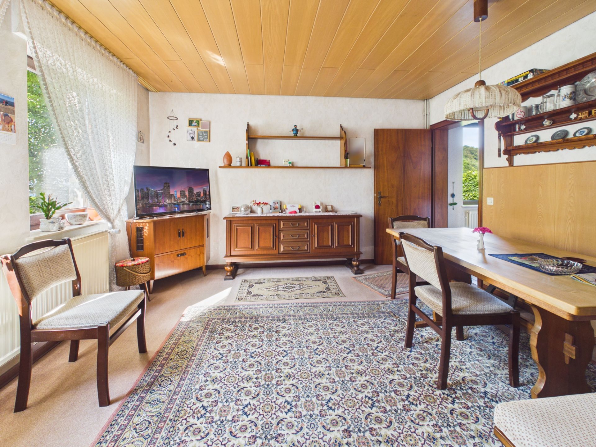 Helles Wohnzimmer mit Teppich, Holzmöbeln, Esstisch, Stühlen und Fernseher am Fenster.
