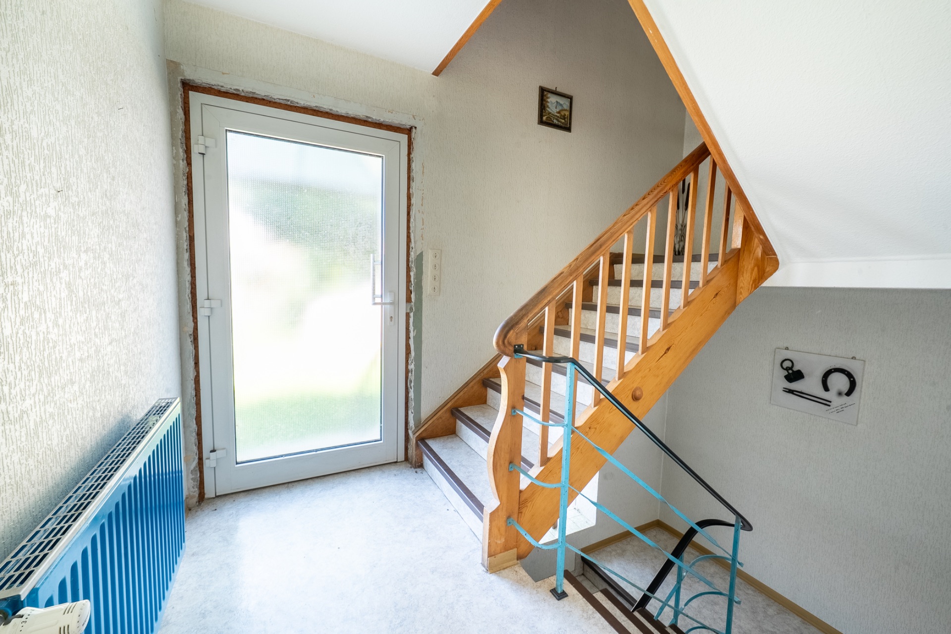 Heller Flur mit Haustür, Treppe mit Holzgeländer und blauem Heizkörper.