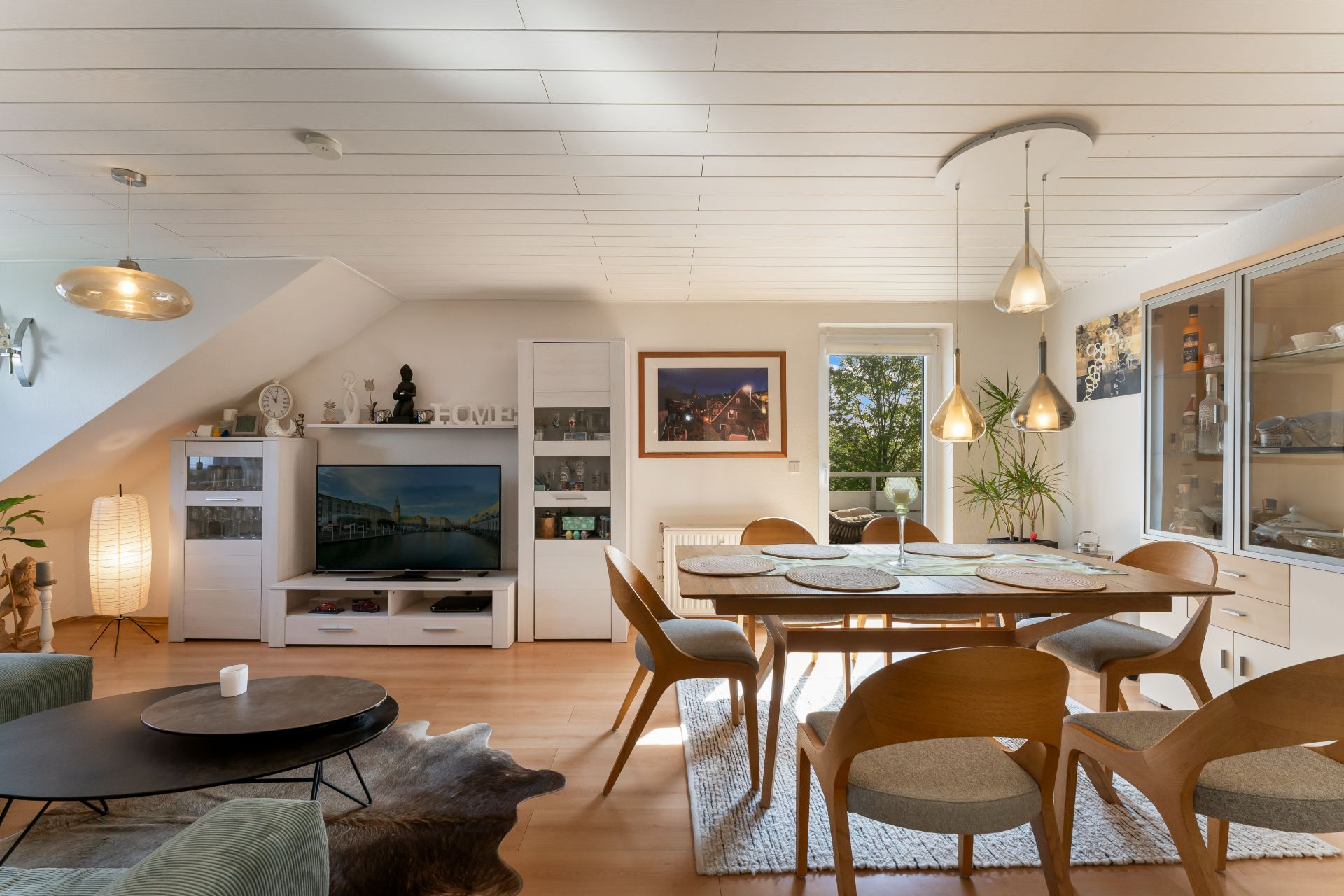 Helles Wohnzimmer mit Esstisch, Stühlen, TV, Vitrine und großen Fenstern.