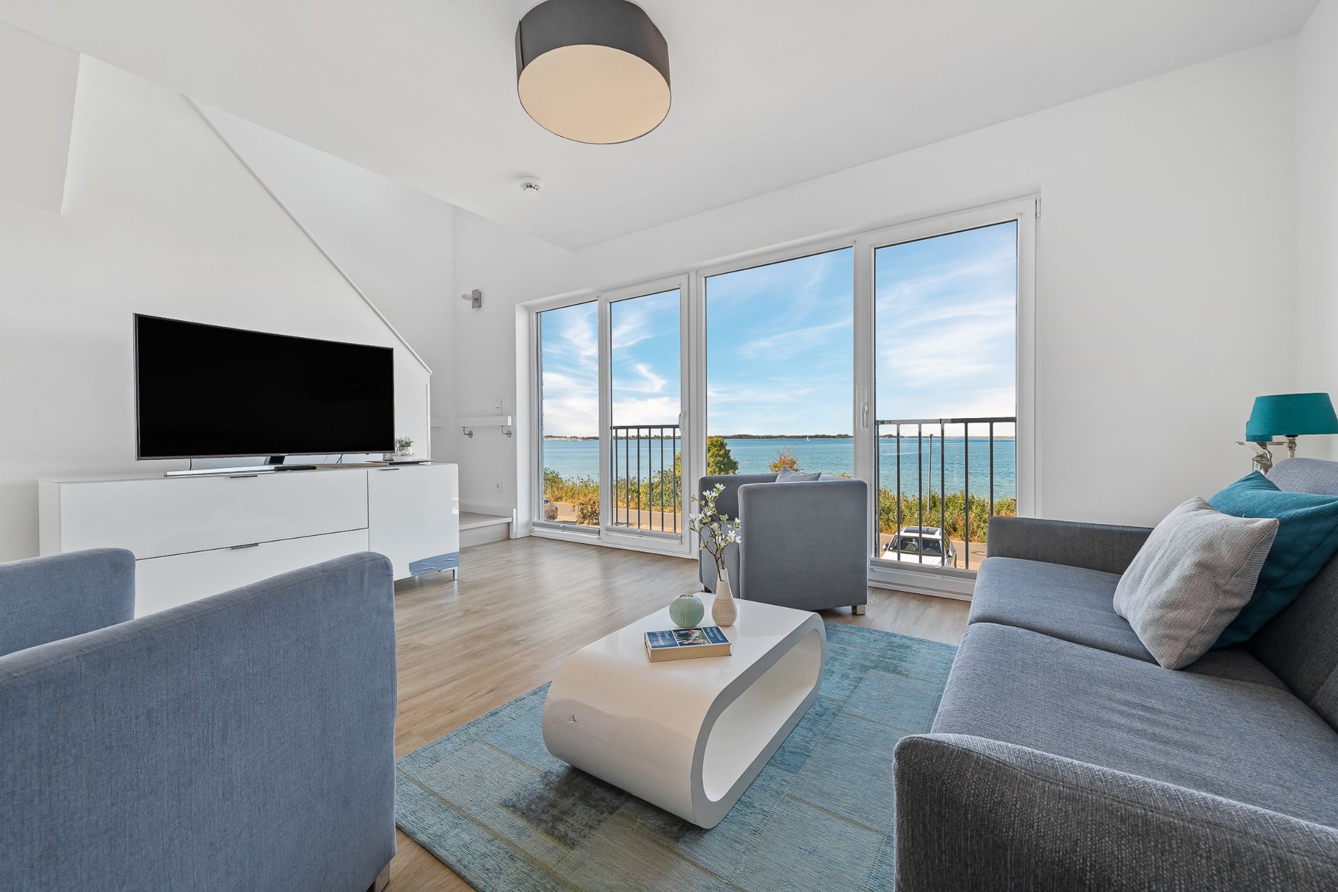 Helles Wohnzimmer mit zwei Sesseln, Sofa, Couchtisch, Fernseher und großem Fenster mit Meerblick.