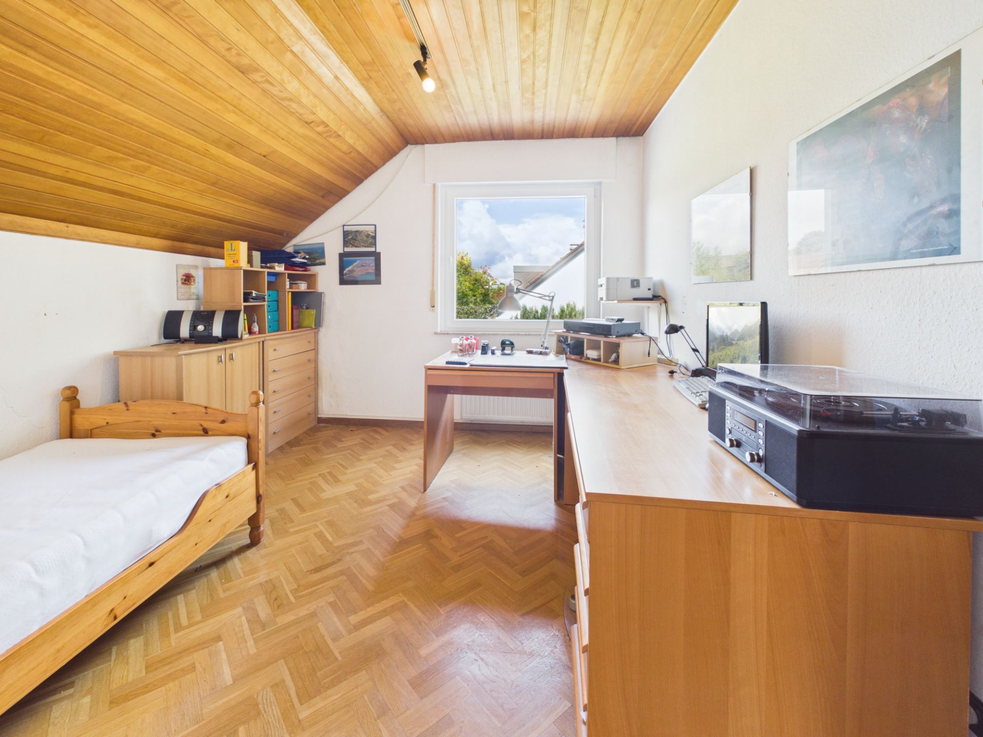 Helles Zimmer mit Holzdecke, Einzelbett, Schreibtisch und Kommode unter einem Fenster.