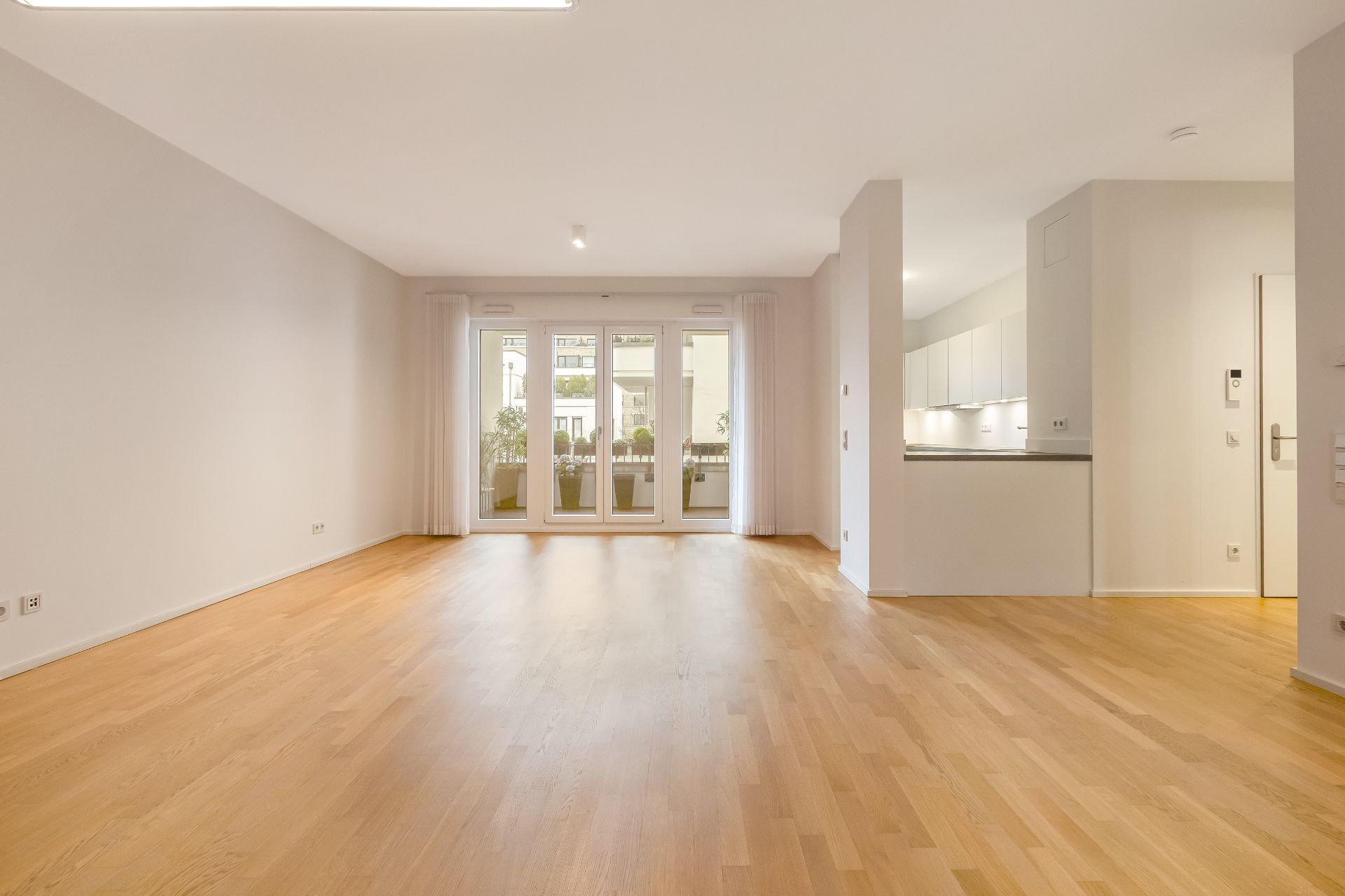 Helles, leeres Wohnzimmer mit Parkettboden, großen Fenstern und offener Küche.