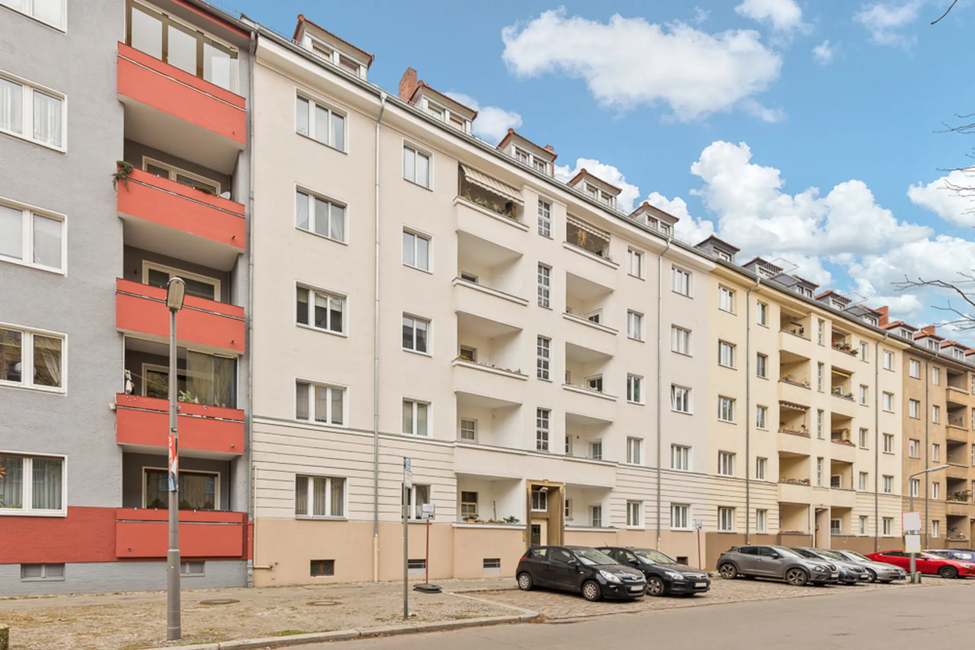 Mehrstöckiges Wohnhaus mit Balkonen, geparkten Autos davor und blauem Himmel im Hintergrund.