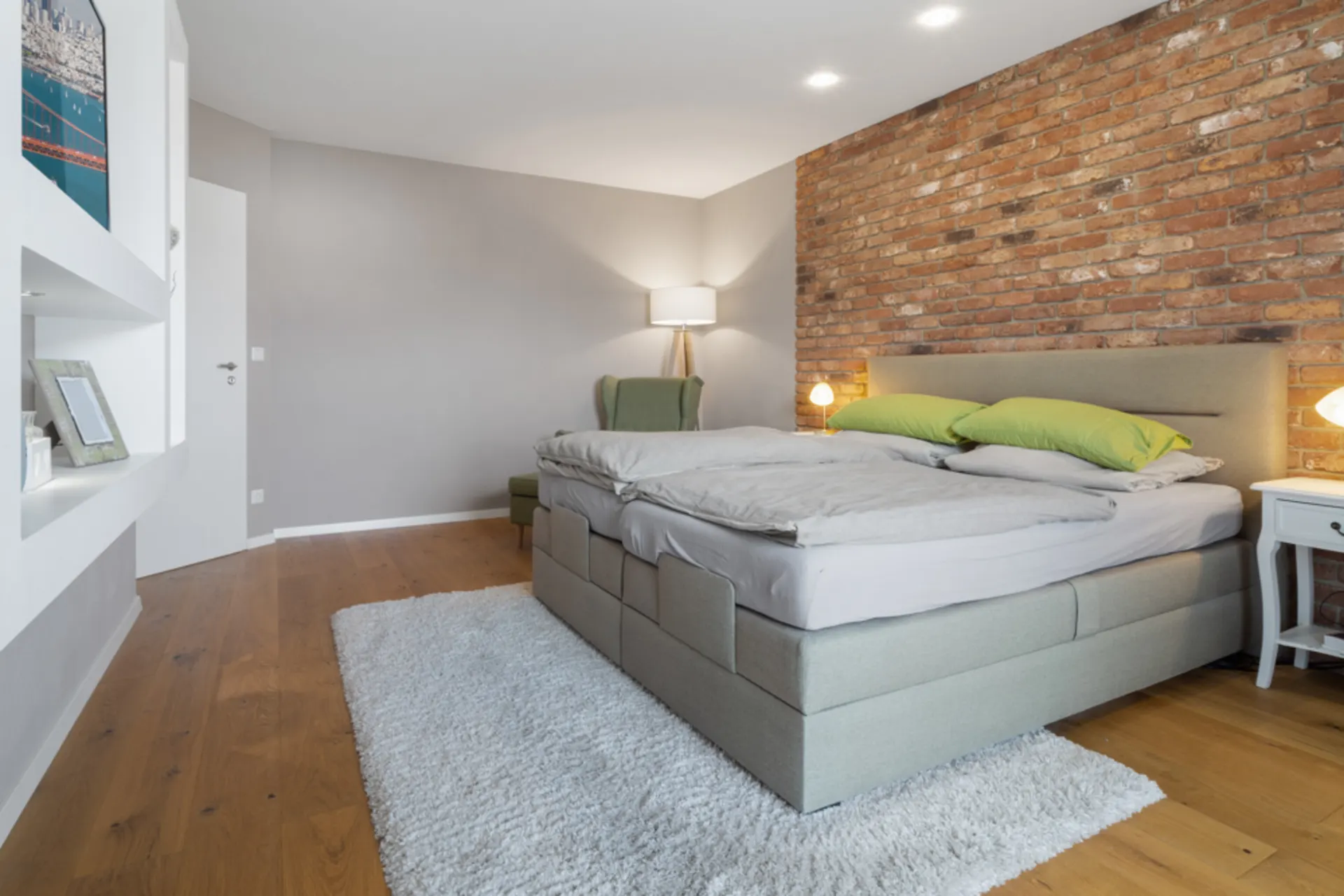 Modernes Schlafzimmer mit Bett, Backsteinwand, Sessel, Nachttischen und Teppich auf Holzboden.