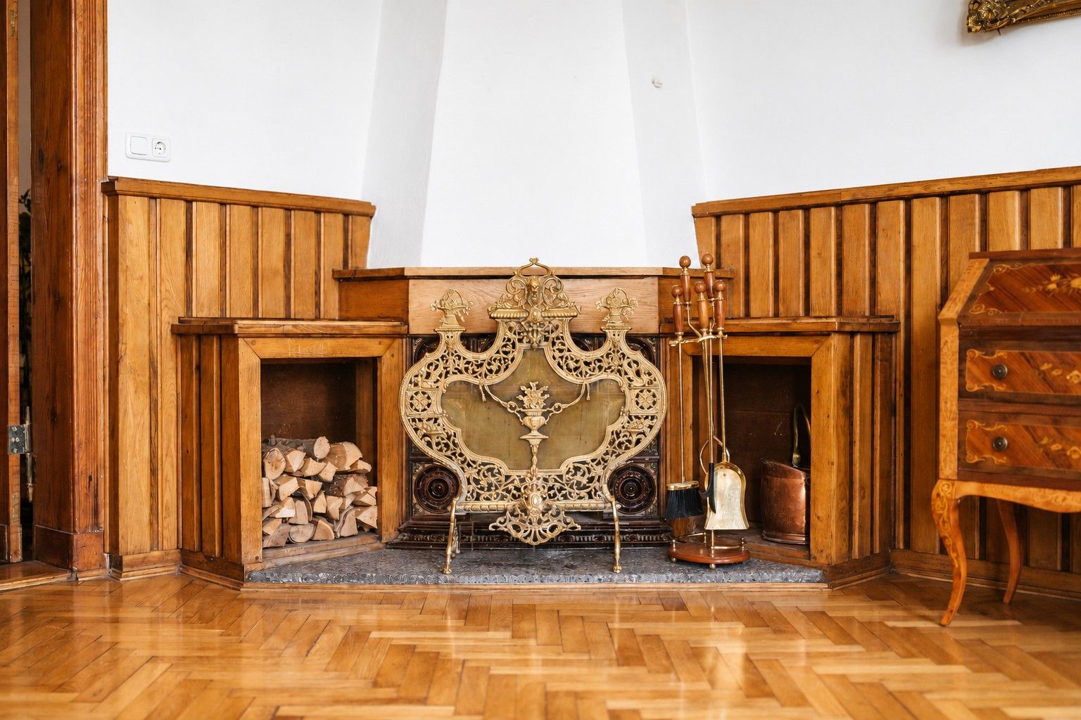 Kamin mit Holzlager, goldenem Kaminaufsatz und Kaminbesteck in einem holzgetäfelten Raum.