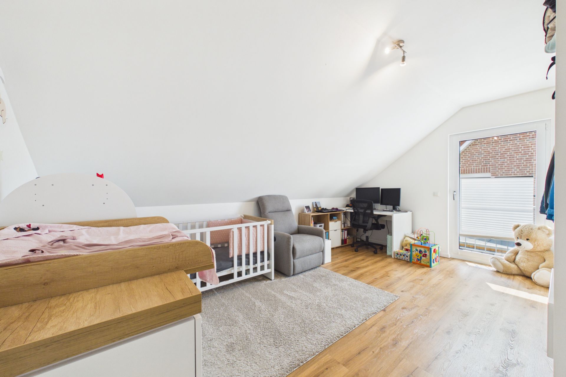 Helles Kinderzimmer mit Wickeltisch, Babybett, Sessel, Schreibtisch und großem Teddybär.
