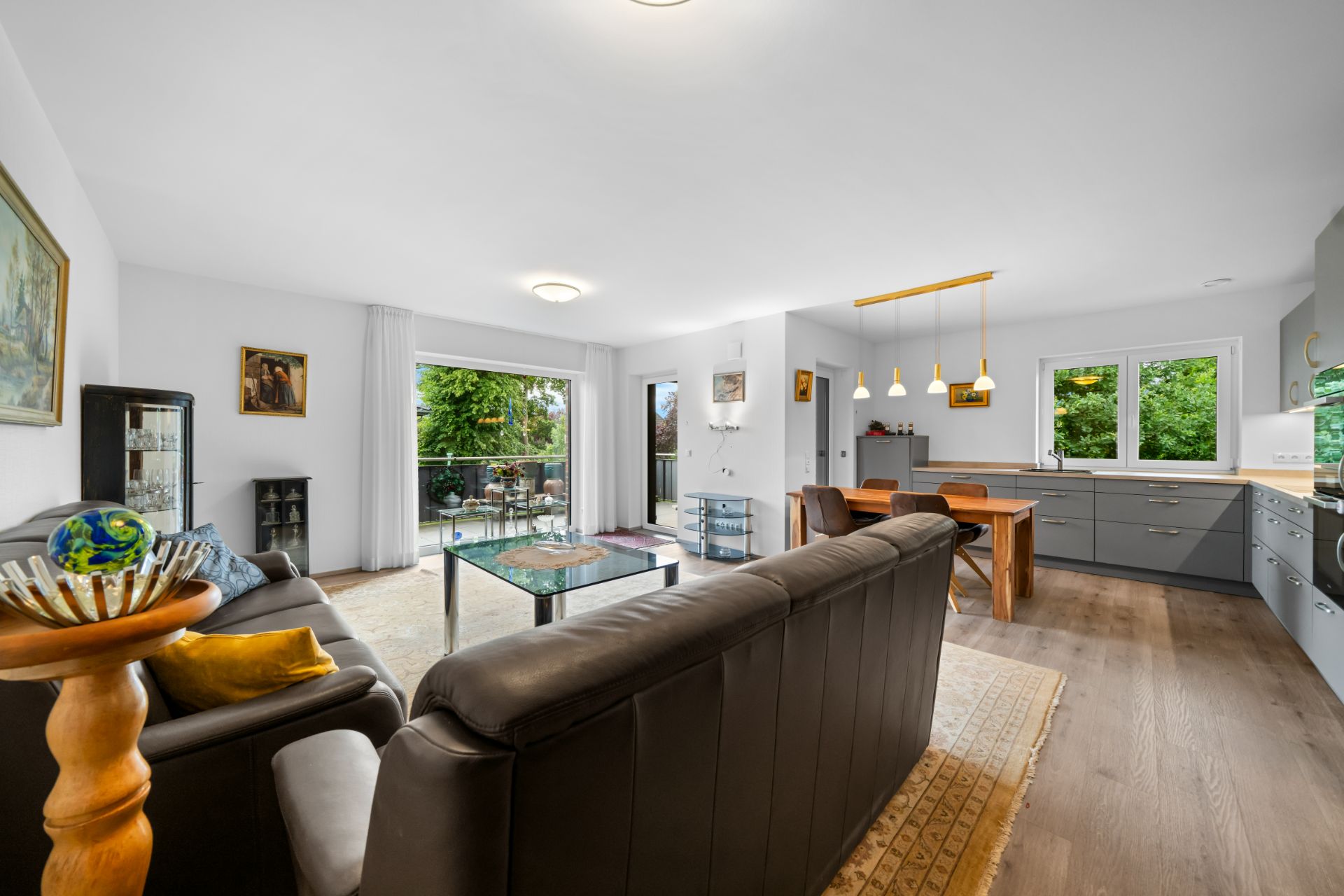 Modernes Wohnzimmer mit offener Küche, Essbereich und Zugang zu einem Balkon mit Blick ins Grüne.