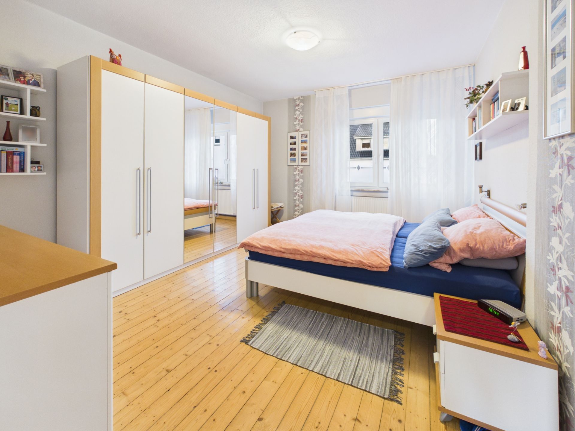 Helles Schlafzimmer mit Holzfußboden, Doppelbett, Kleiderschrank und Fenstern mit weißen Vorhängen.