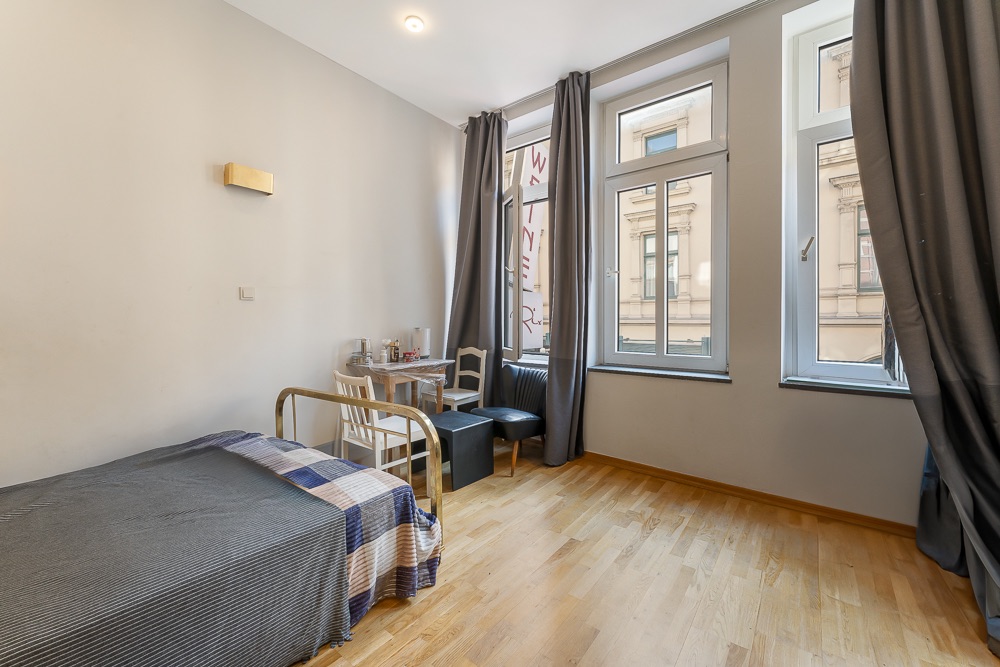 Helles Zimmer mit Einzelbett, kleinem Esstisch und drei großen Fenstern mit Vorhängen.