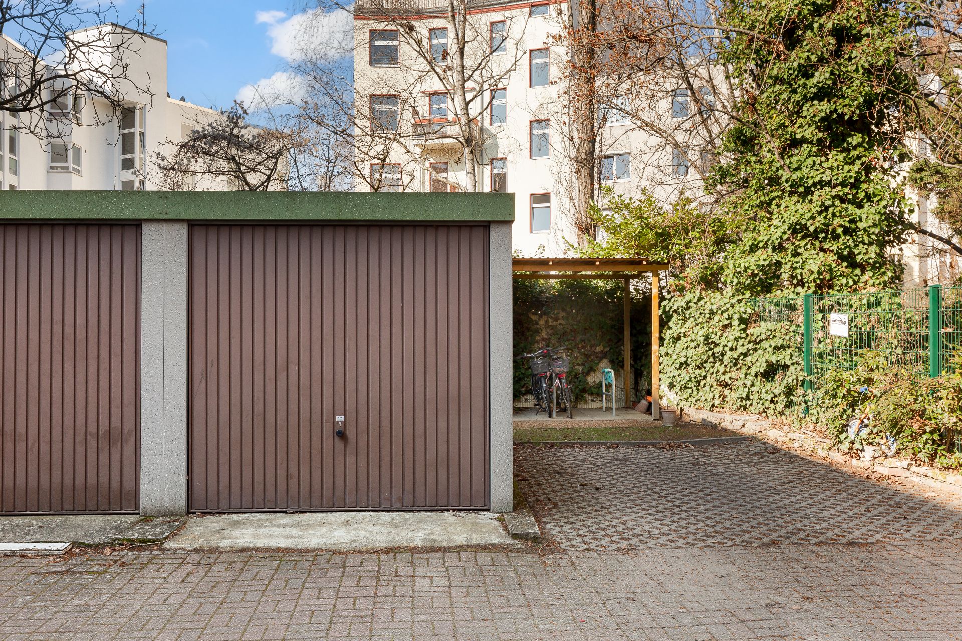 Garage mit Holztor und überdachtem Fahrradstellplatz daneben auf einem gepflasterten Hof.