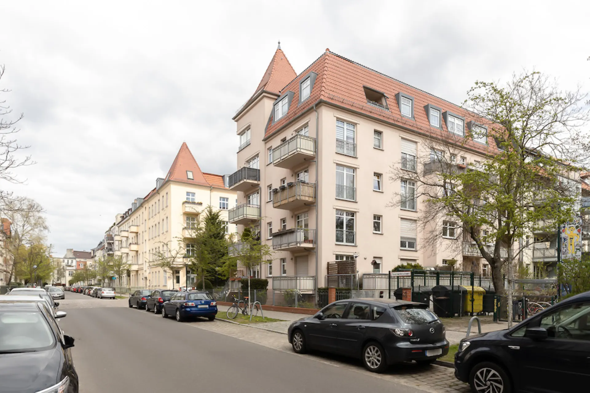 Mehrstöckiges Wohnhaus mit Balkonen an einer ruhigen Straße, geparkte Autos und Bäume am Straßenrand.